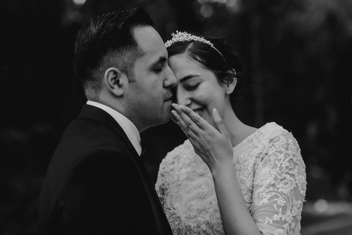 Black and white photo of groom kissing blushing bride on cheek, bride covering smile with hand.