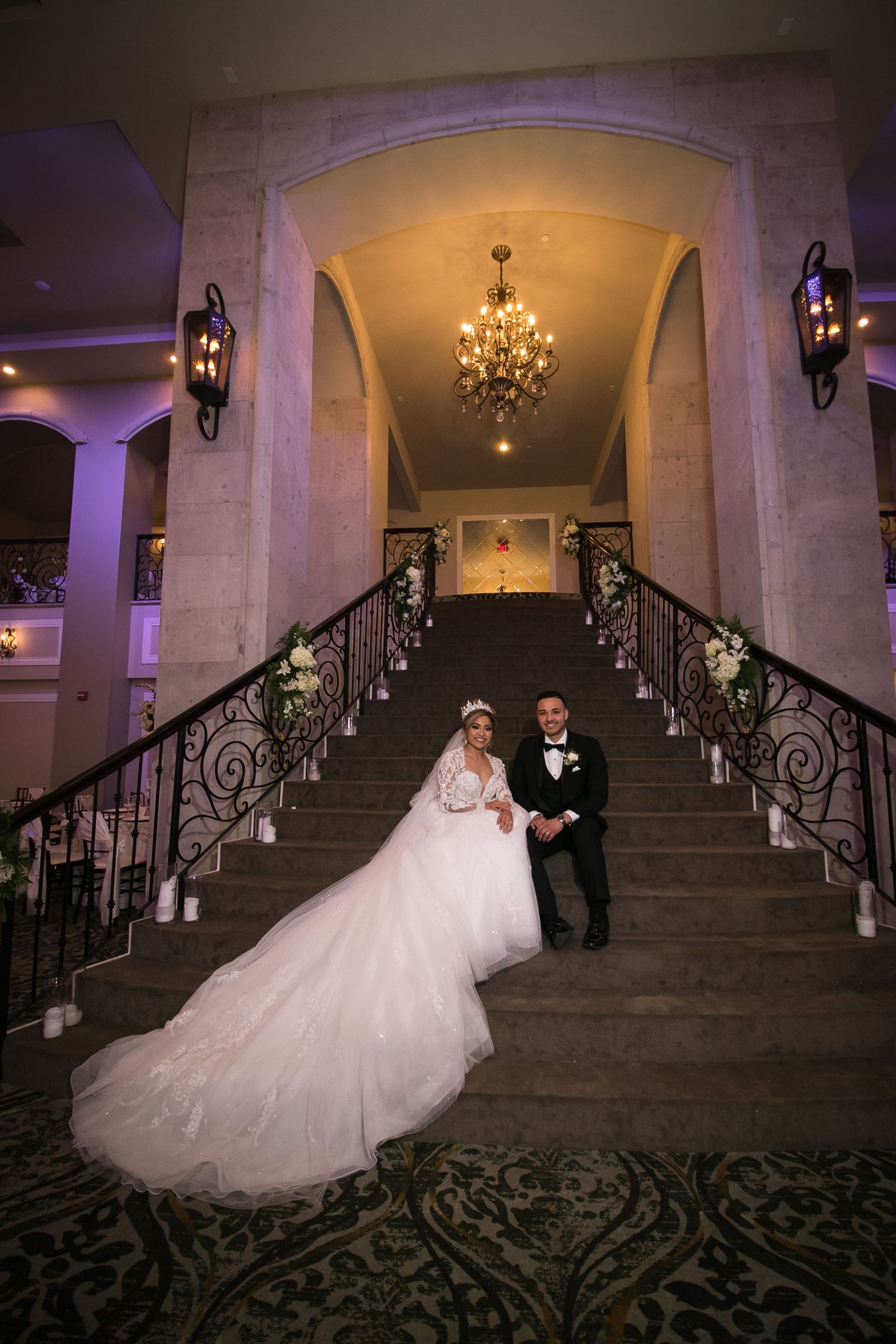 Bride and groom pose on elegant grand staircase with iron railings and chandelier at wedding venue.