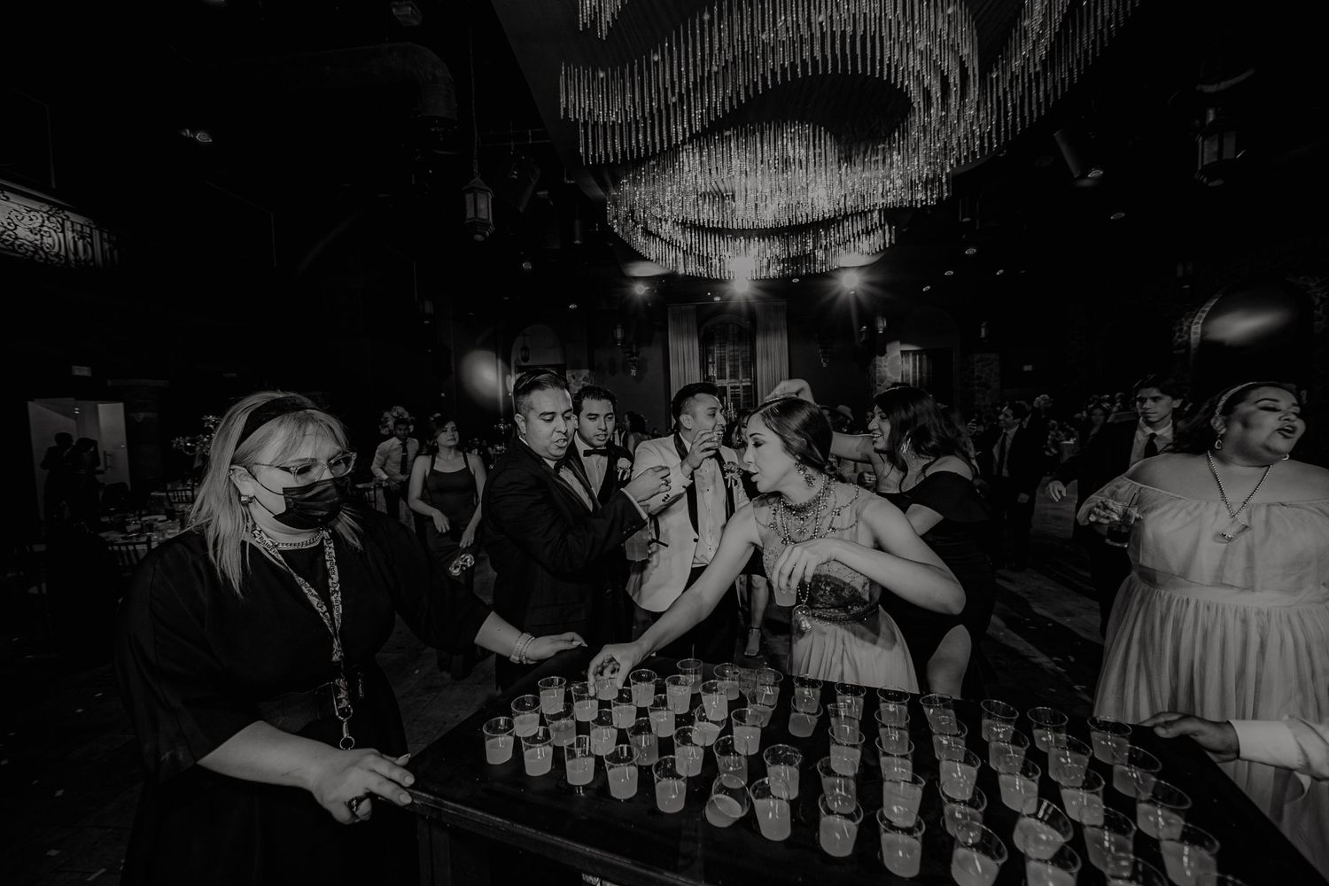 Black and white photo of wedding guests gathering around a bar with shot glasses at an elegant reception venue.