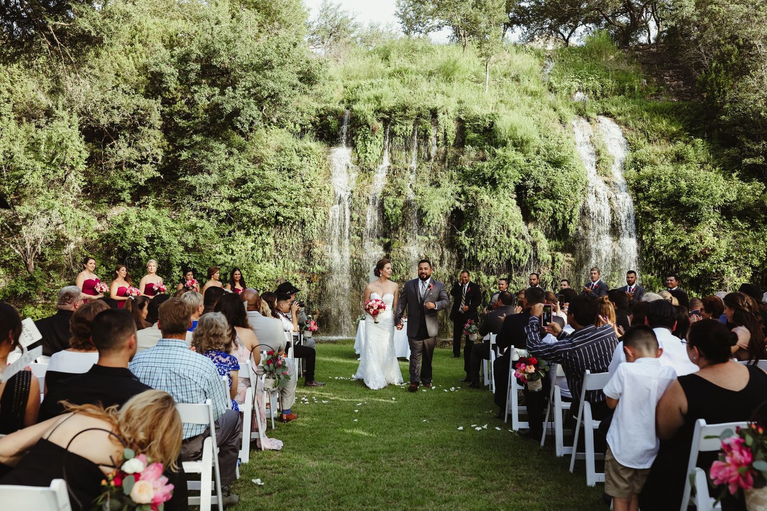 Couple walks down outdoor wedding aisle surrounded by guests with a stunning waterfall backdrop.