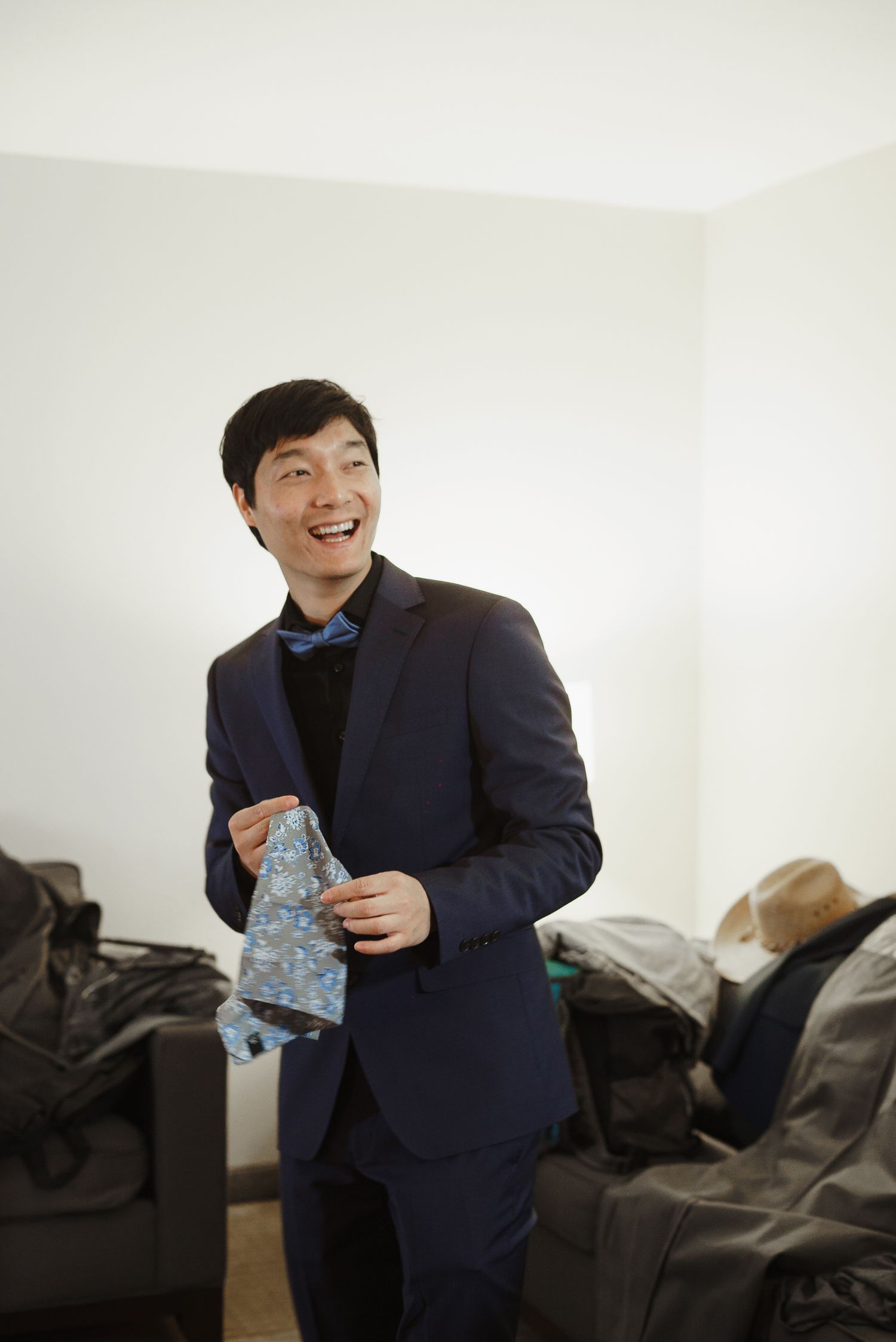 Smiling groom in navy suit holding blue floral tie while getting ready indoors.
