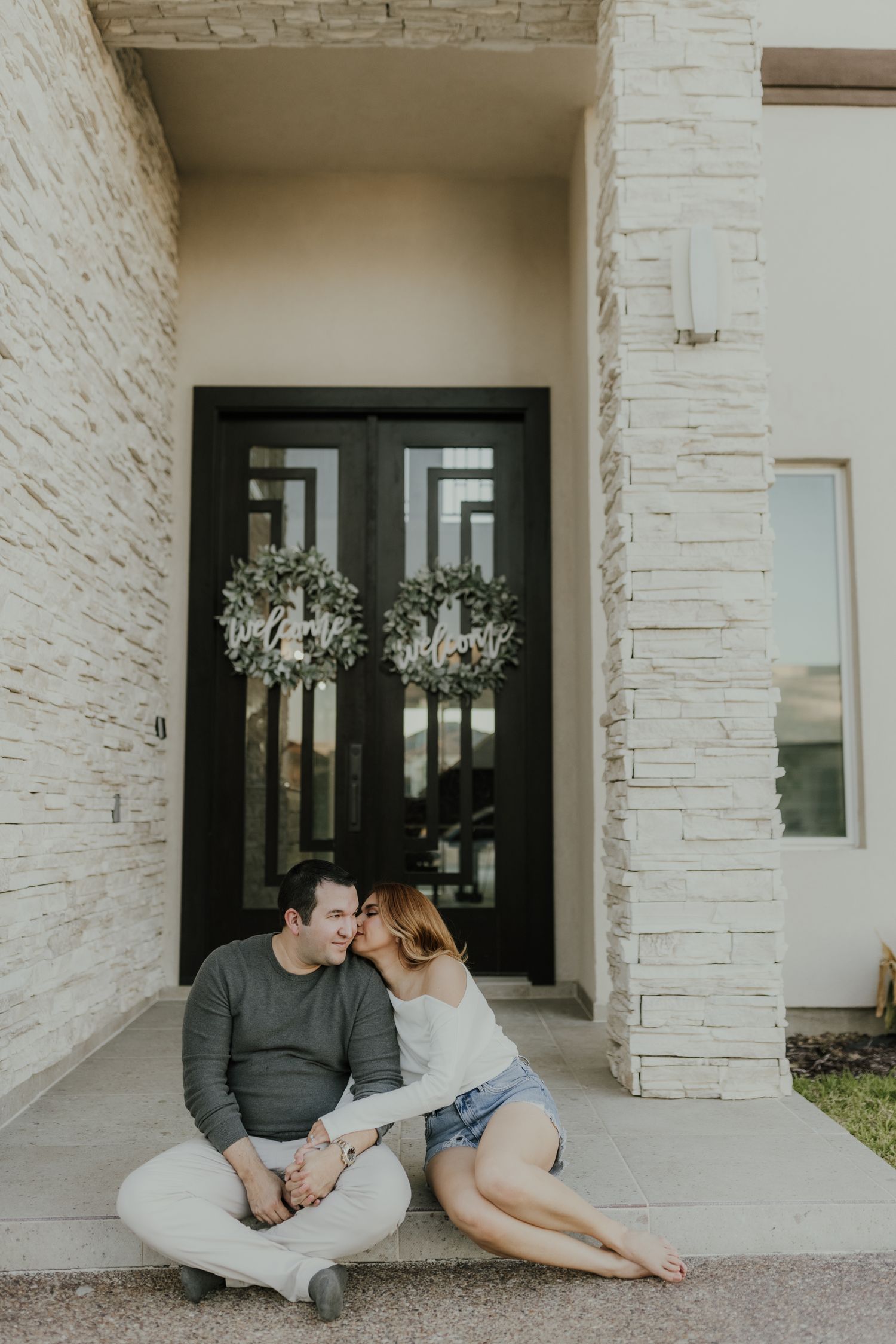 Couple sitting on front porch steps sharing a tender moment, with decorative wreaths on black double doors behind them.
