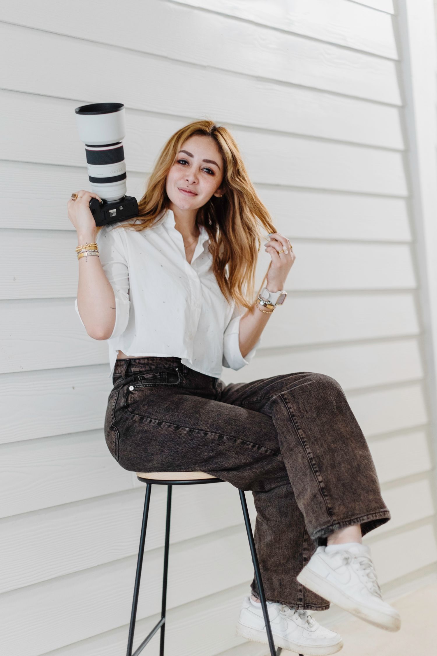 Professional photographer in white shirt sits on stool with camera against white wall background.