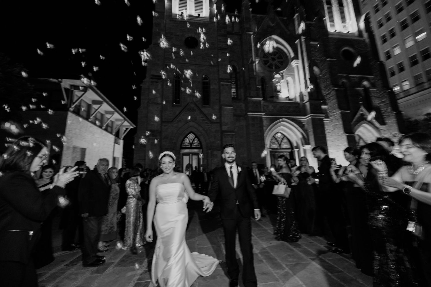 Black and white photo of newlyweds exiting a church at night amid confetti, surrounded by cheering guests.