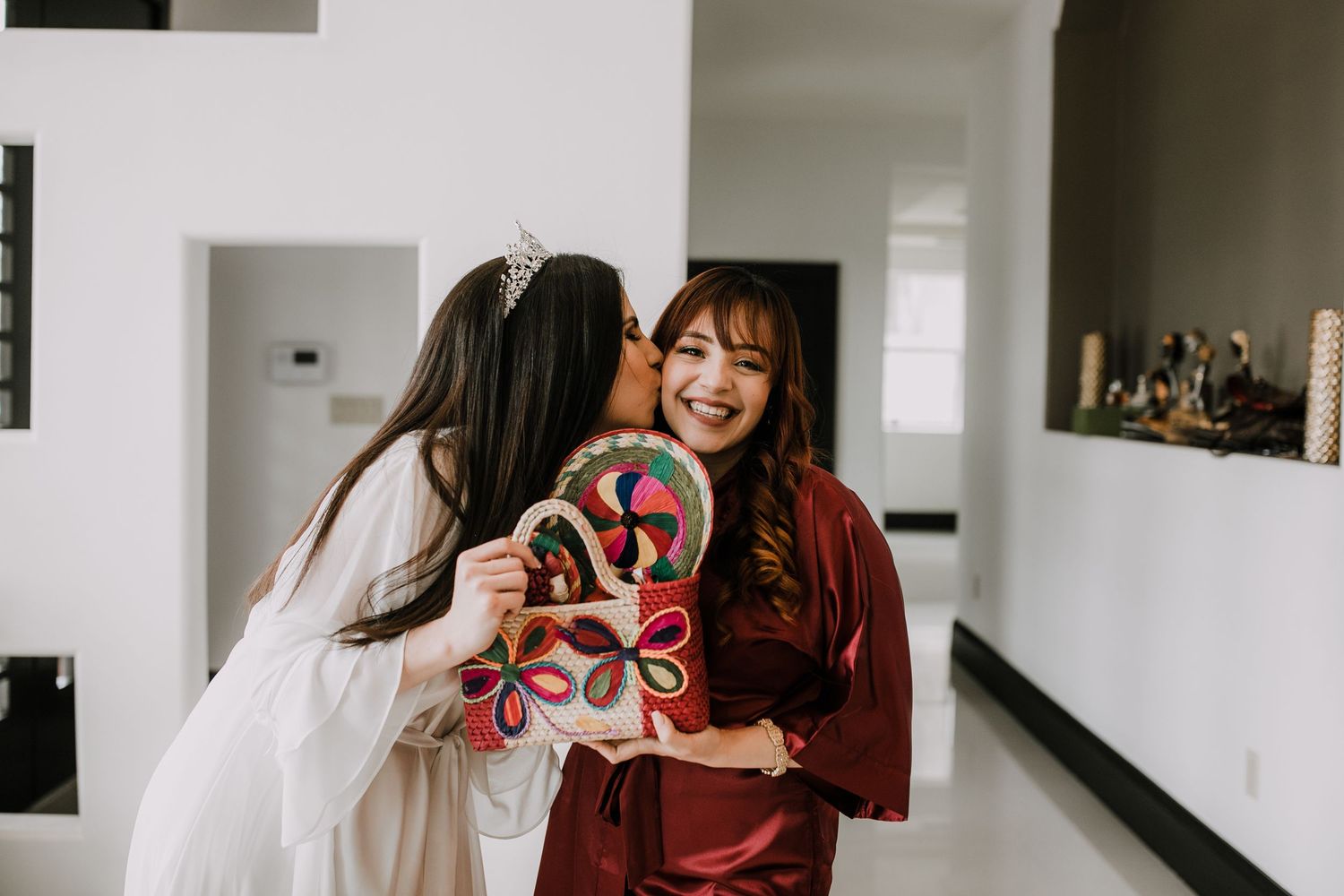 Bride in white robe kisses bridesmaid on cheek while she laughs holding a colorful decorative fan.