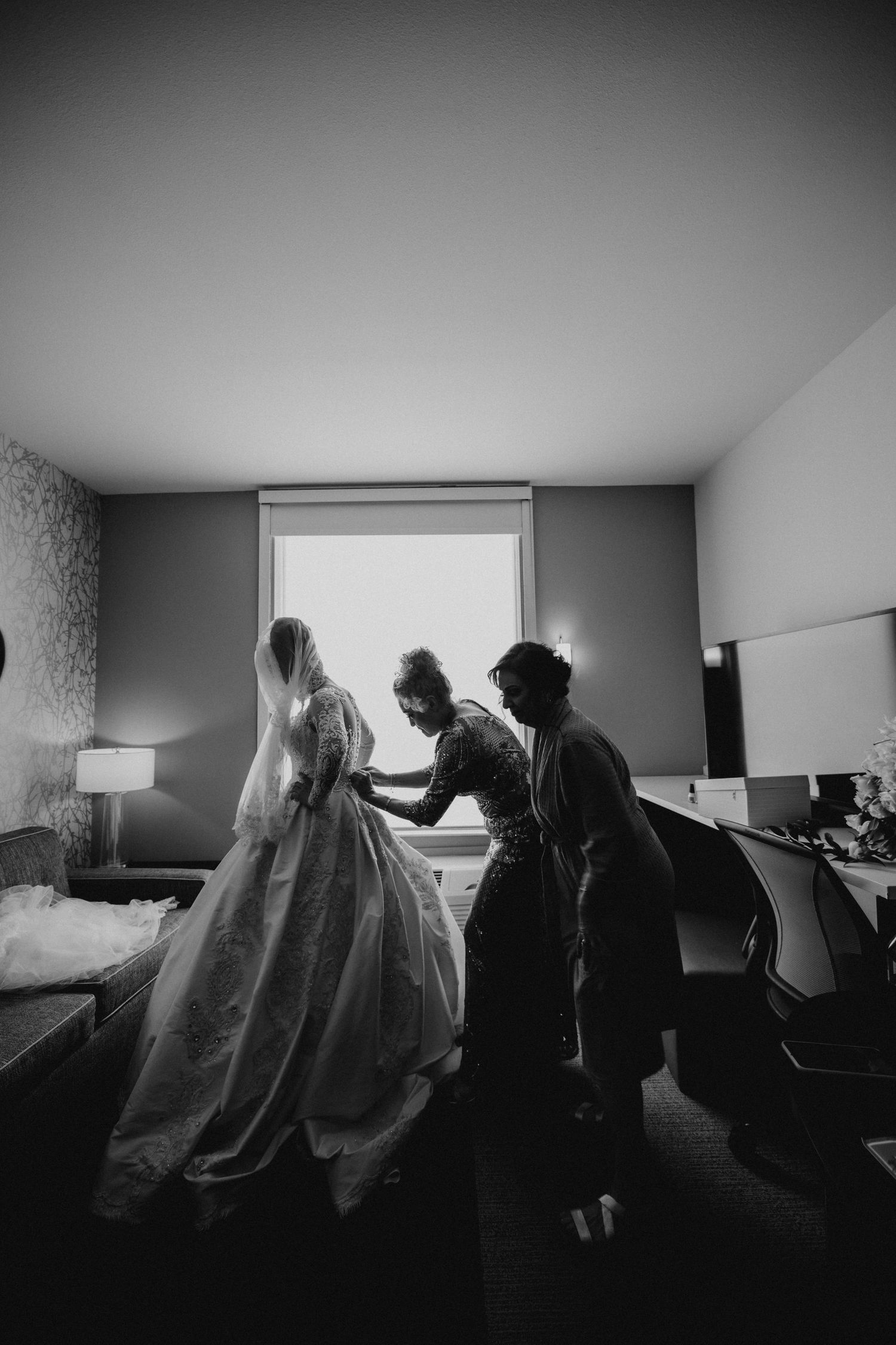 Black and white photo of bride in ballgown being assisted by two helpers in hotel room.