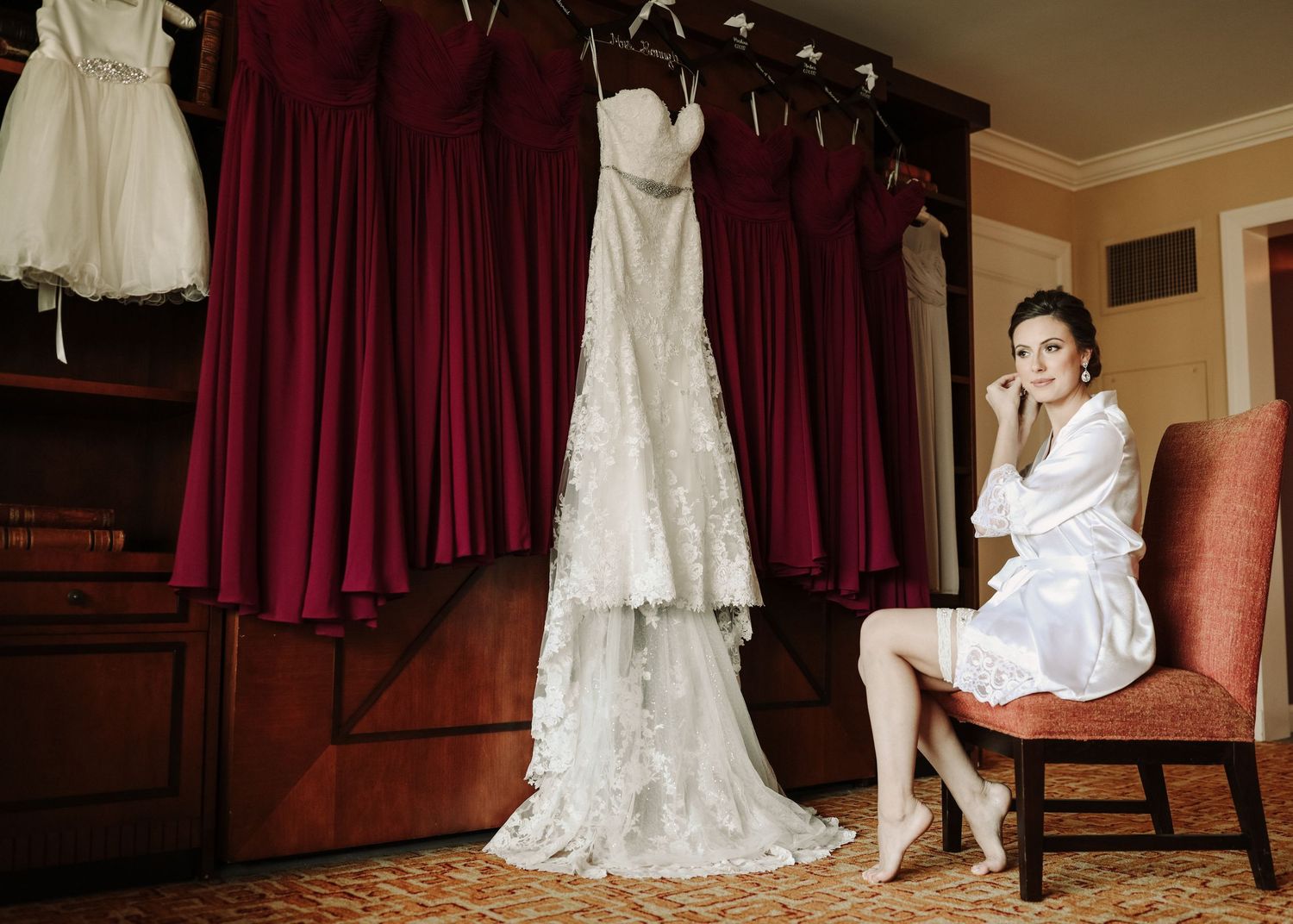 Bride in white robe sits contemplatively beside her lace wedding gown in bridal suite.