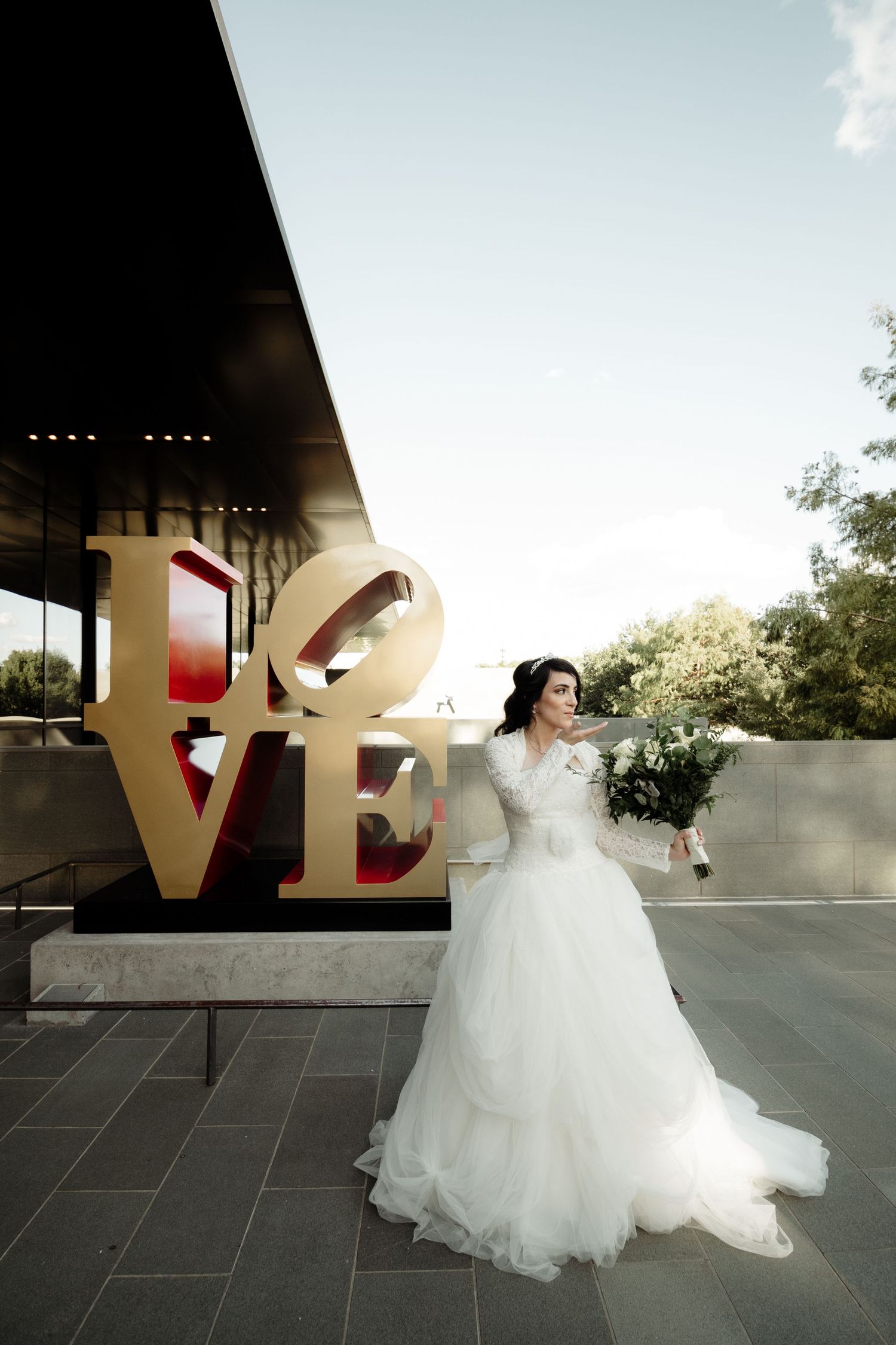 Elegant bride in white ball gown poses beside the iconic LOVE sculpture outdoors on her wedding day.