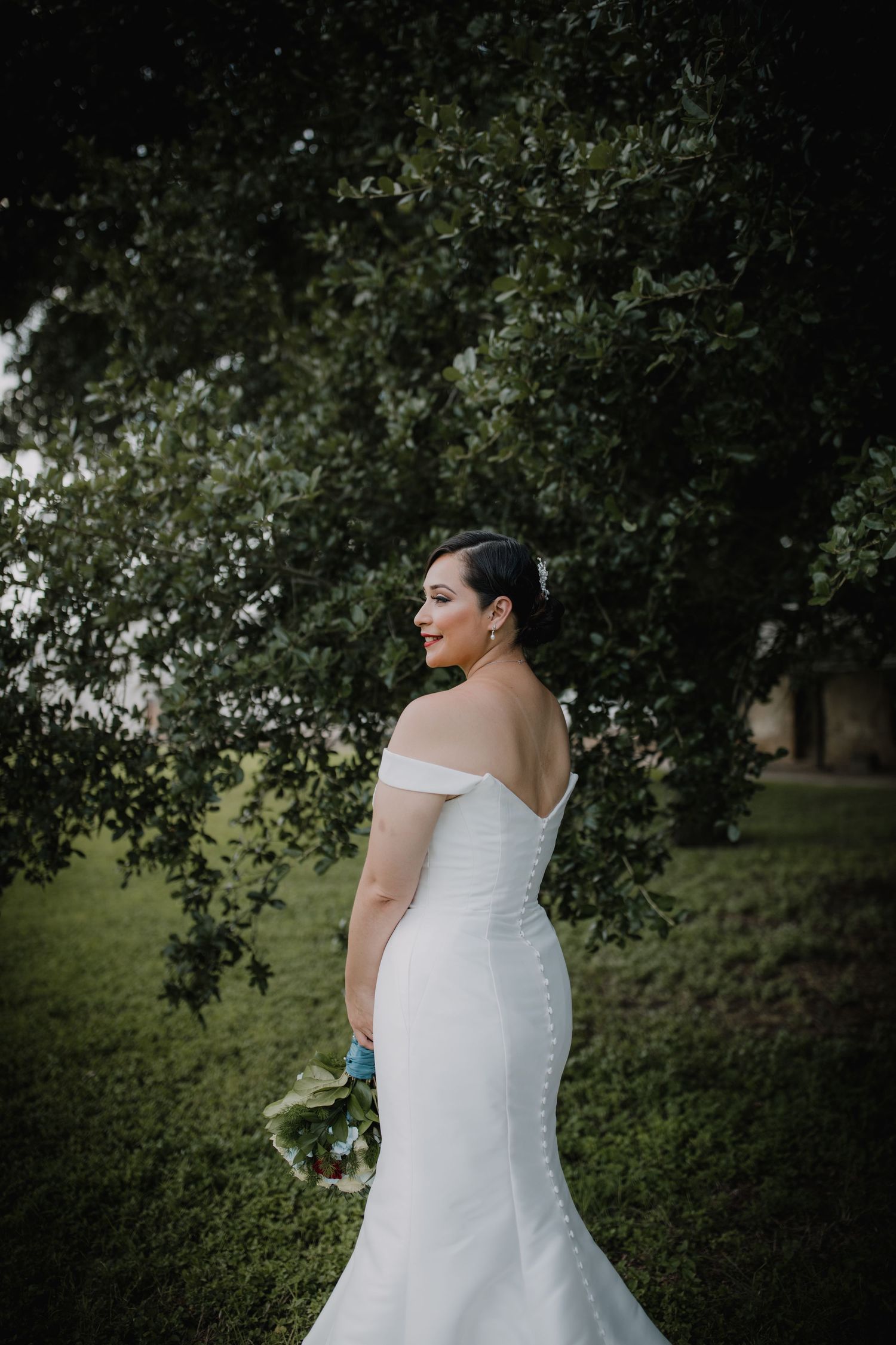 Bride in off-shoulder mermaid wedding gown holds bouquet while standing before lush green trees.
