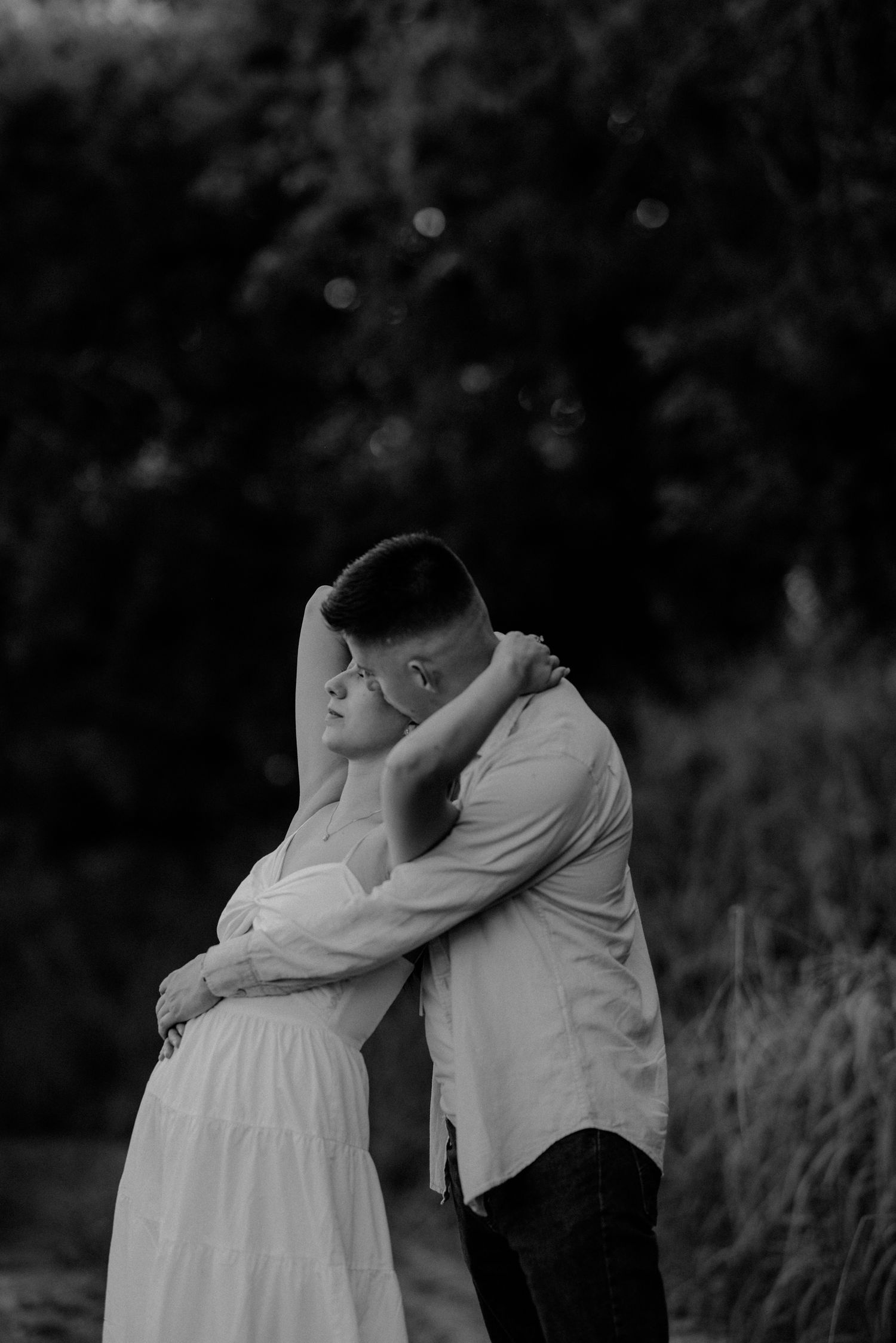 Black and white photo of a couple embracing outdoors, woman in white dress leaning back as man holds her close.