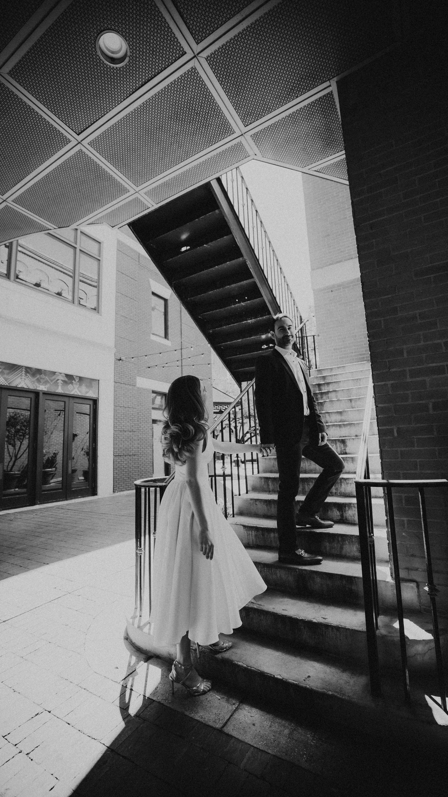 Black and white photo of a bride and groom walking up outdoor stairs at a modern building venue.
