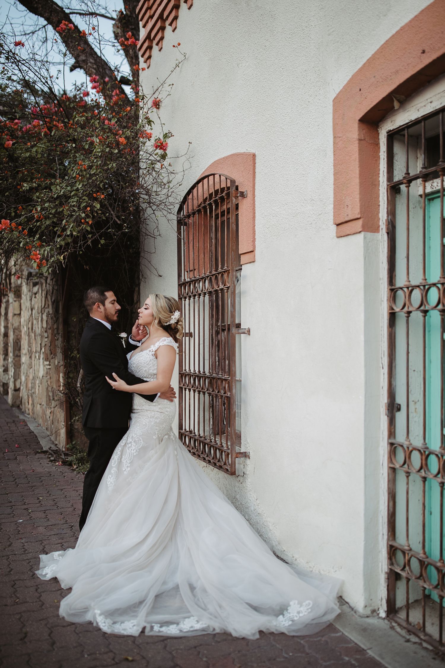 Bride and groom share a romantic moment in a charming alley with colorful flowers and teal doors.
