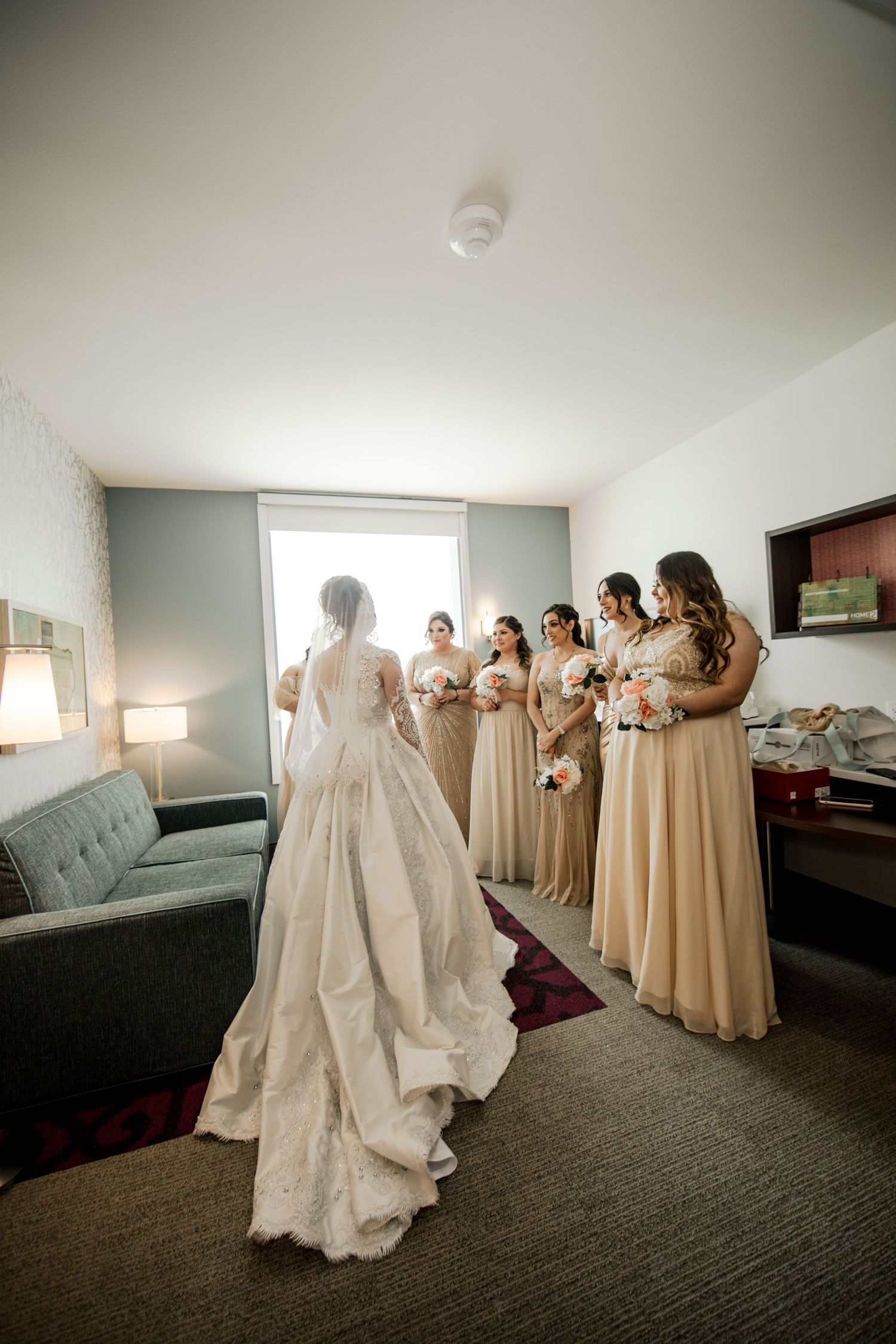 Bride in elegant lace gown with long veil faces bridesmaids in champagne dresses holding bouquets in hotel room.