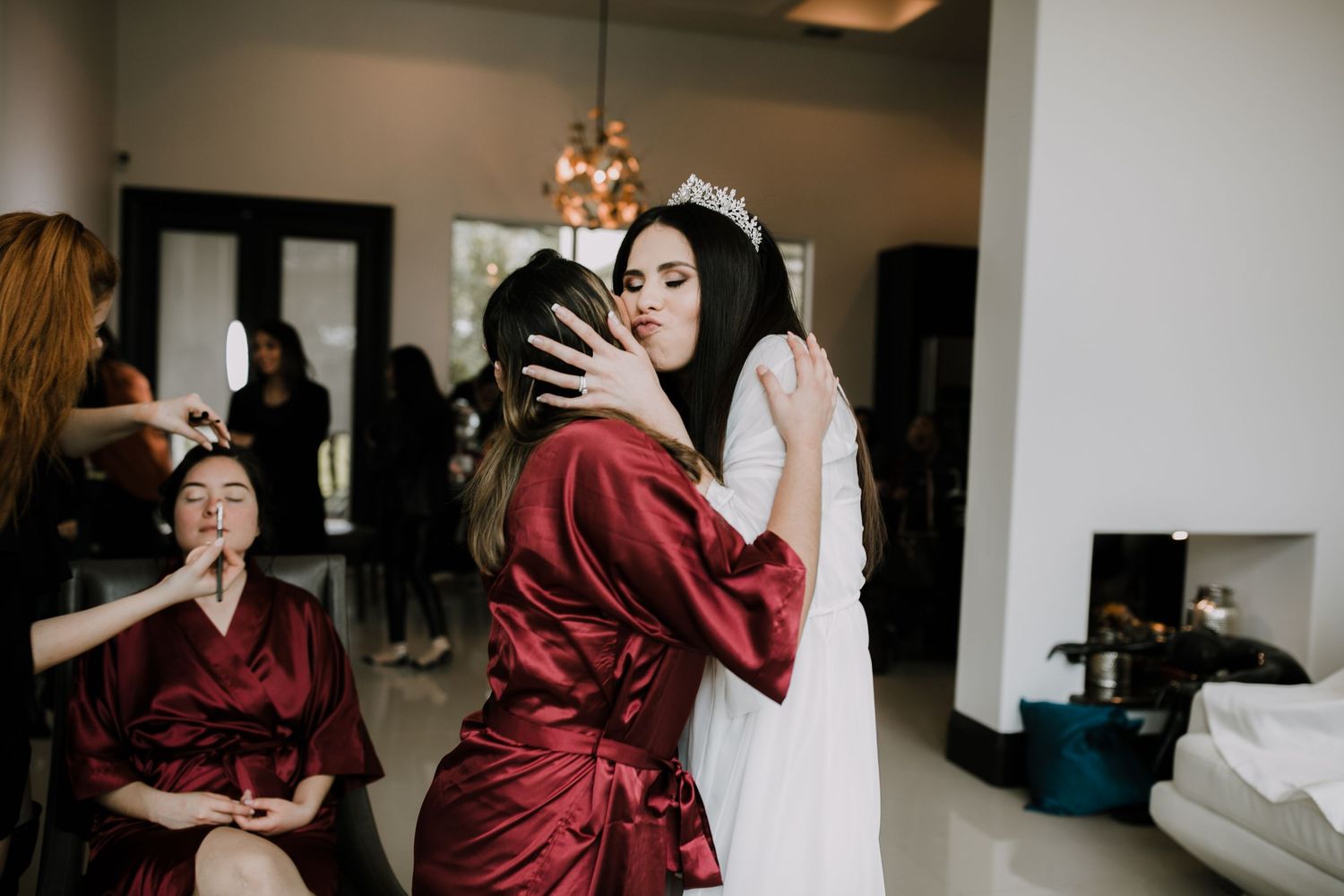 Bride wearing a tiara embracing a bridesmaid in a red robe during wedding preparations.