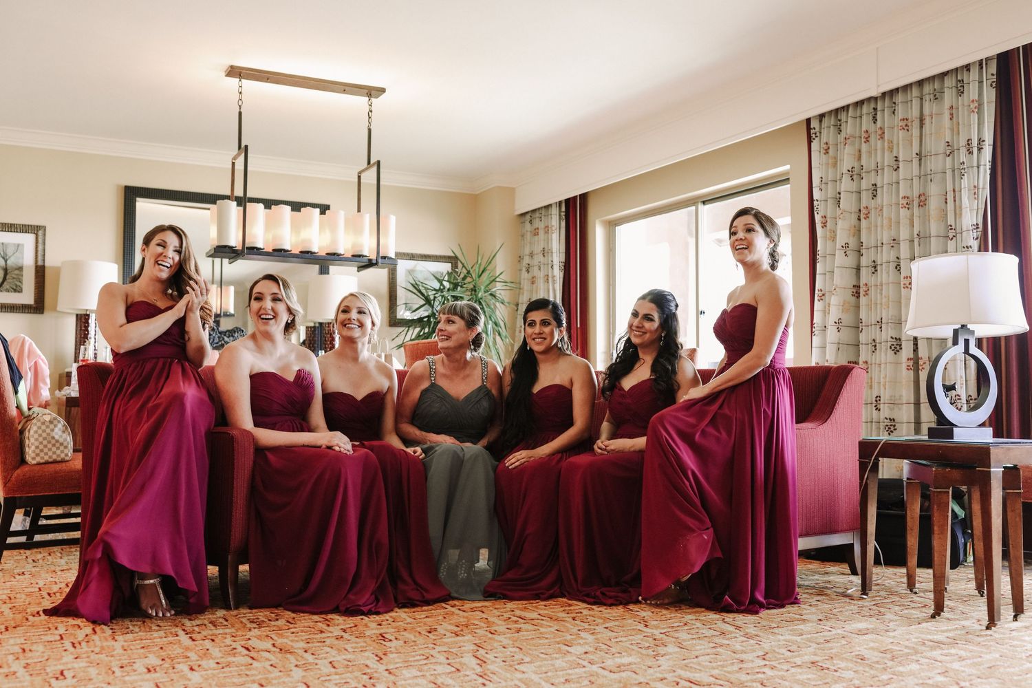 Bridesmaids in burgundy gowns pose together in a warmly lit hotel suite before the wedding.