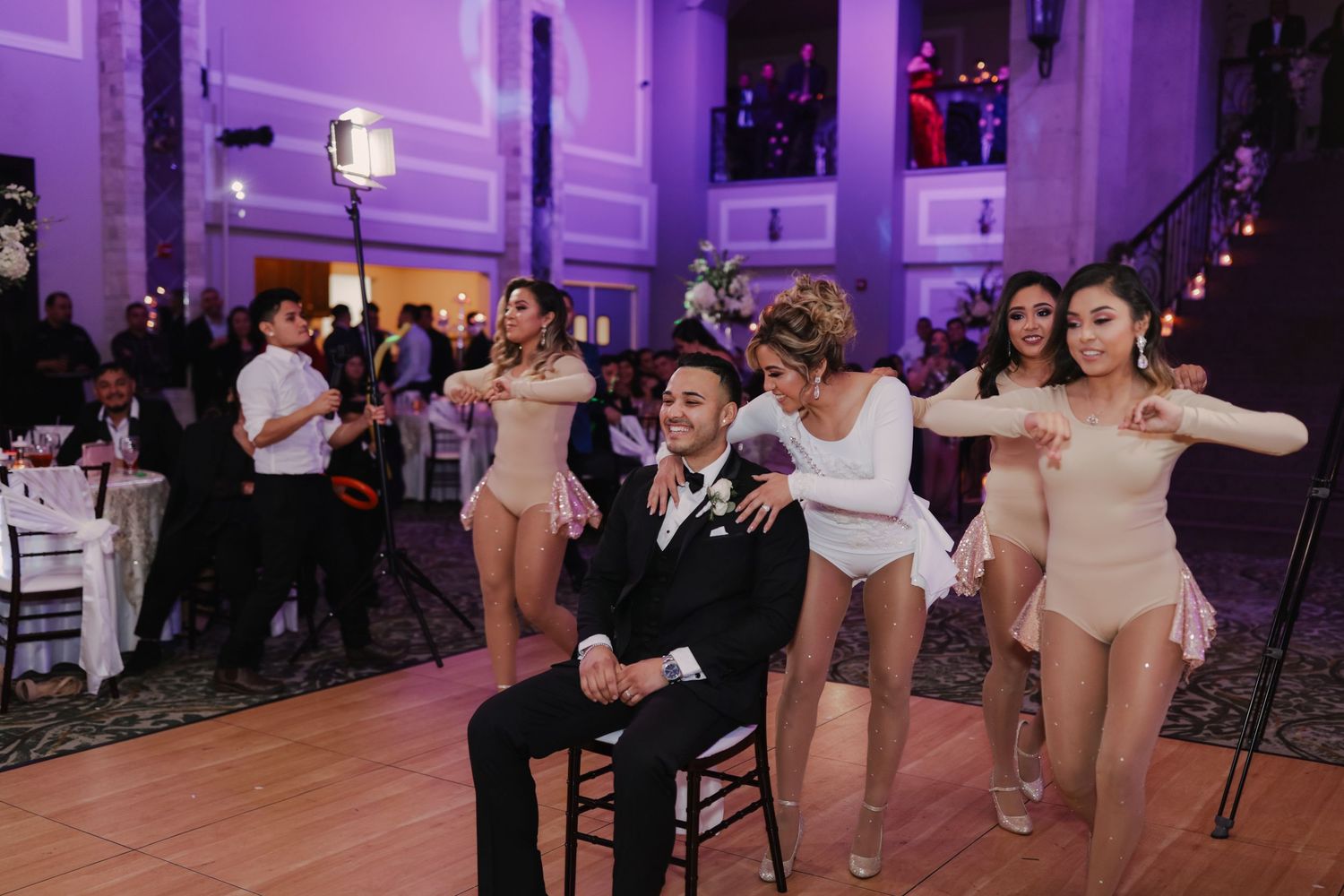 Groom sits on chair smiling as dancers in nude bodysuits perform around him at wedding reception.