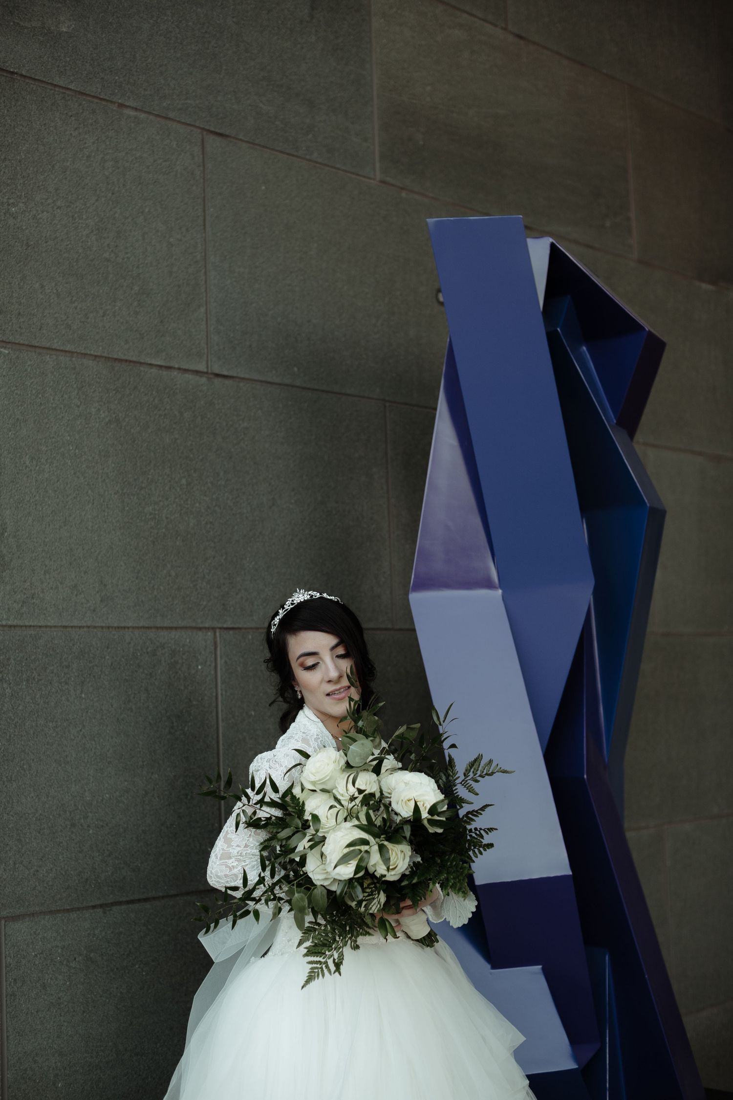 Bride in white ballgown holding lush white rose bouquet, standing beside blue geometric sculpture against stone wall.