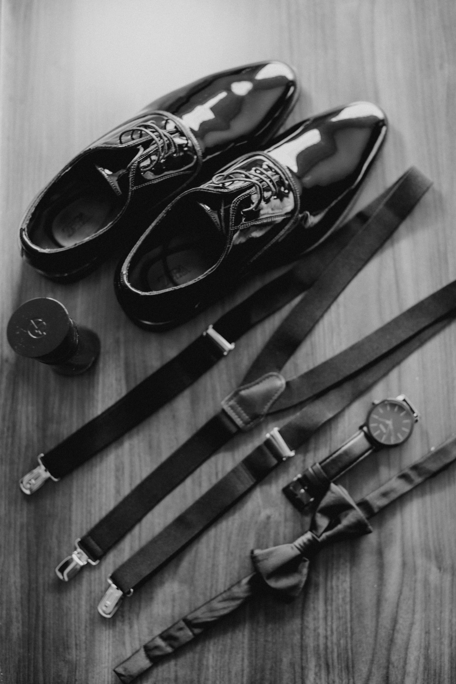 Polished black oxford shoes with suspenders, bow tie, watch, and cufflinks arranged on wooden surface.