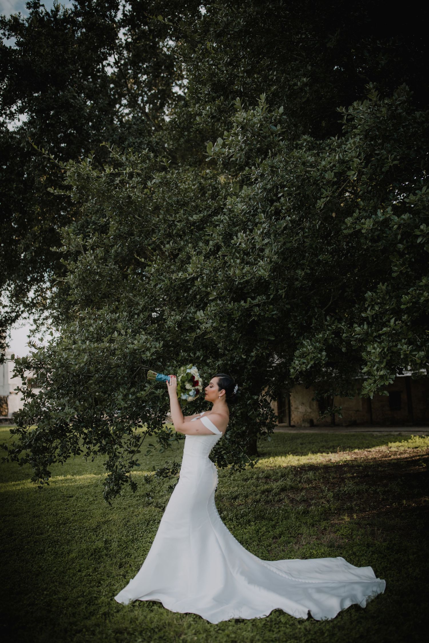 Bride in a sleek white gown with a long train holds her bouquet under a large oak tree outdoors.