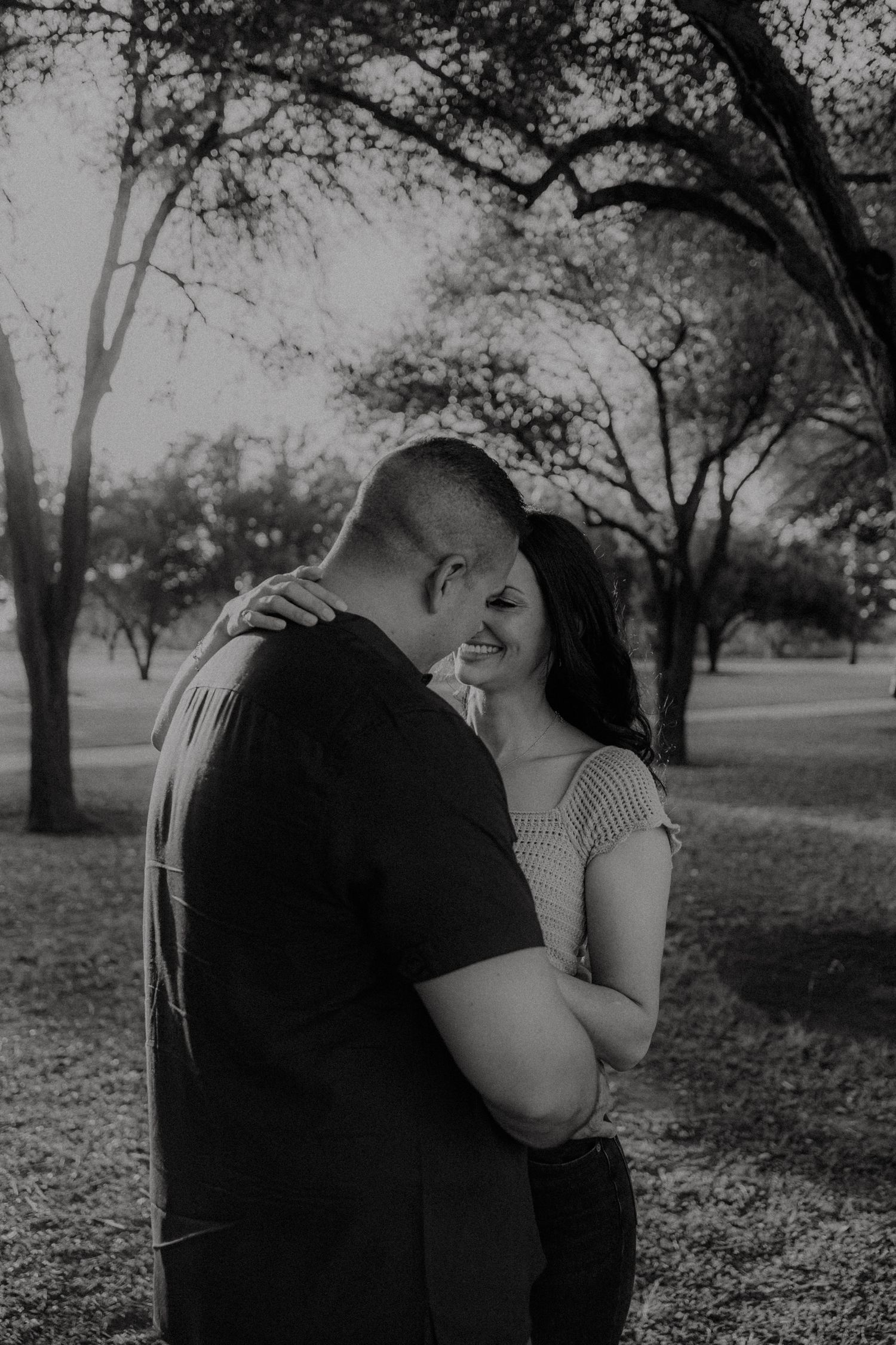 Black and white photo of a couple embracing and smiling in a park with bare trees in the background.