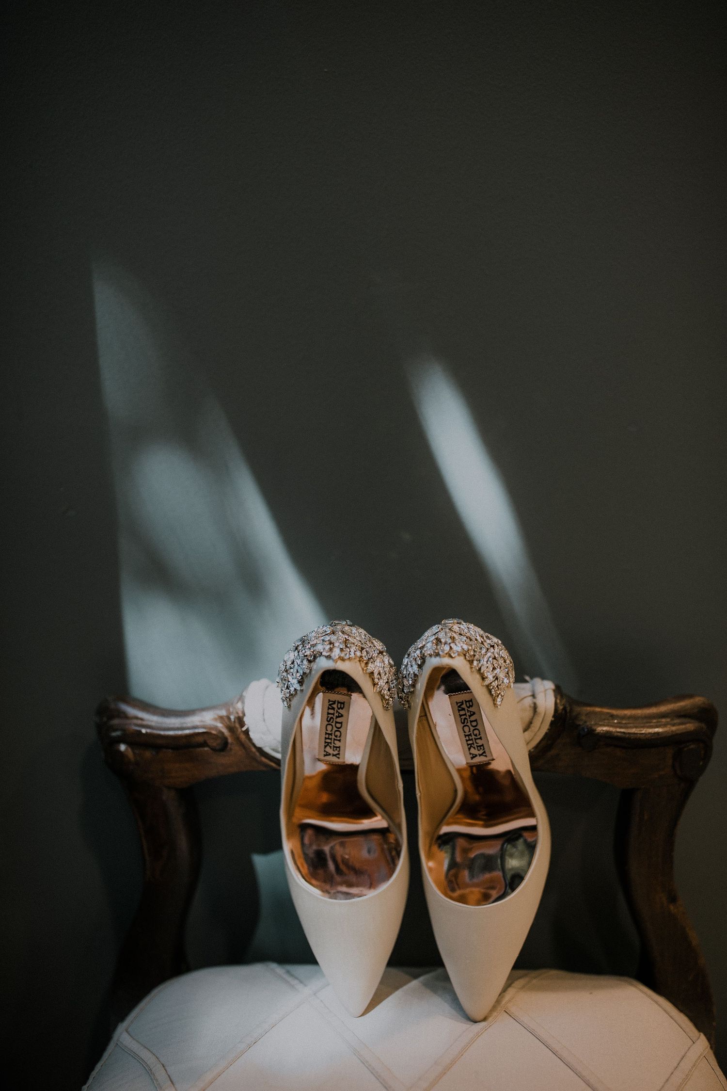 White Badgley Mischka bridal heels with crystal embellishment displayed on vintage wooden chair against gray wall.