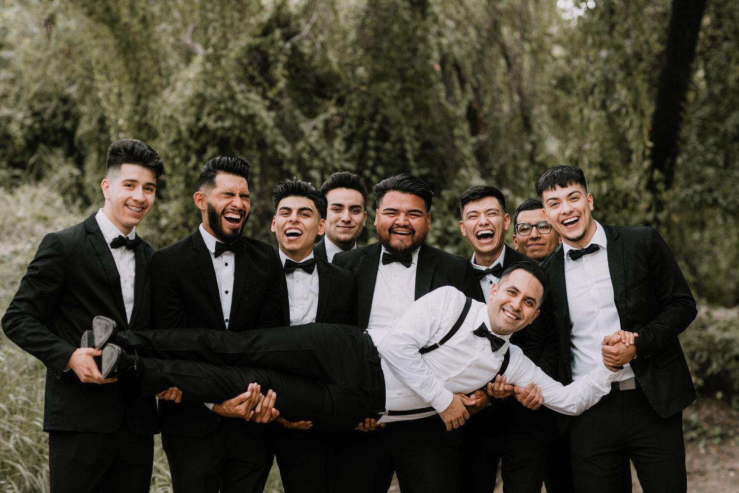 Groomsmen in black tuxedos lifting groom horizontally while laughing outdoors.