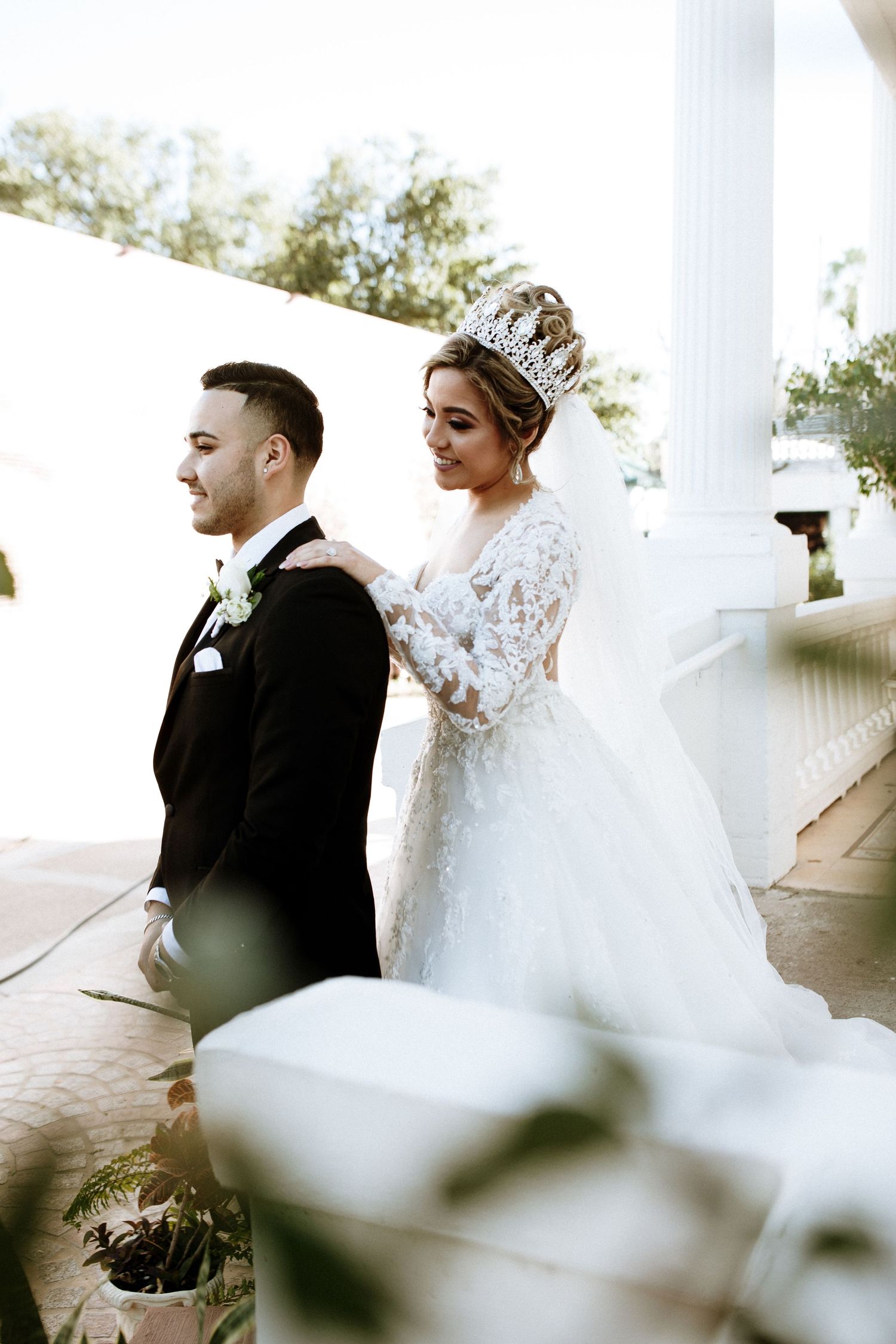 Bride in crown and lace gown stands behind groom in tuxedo during wedding first look outdoors.
