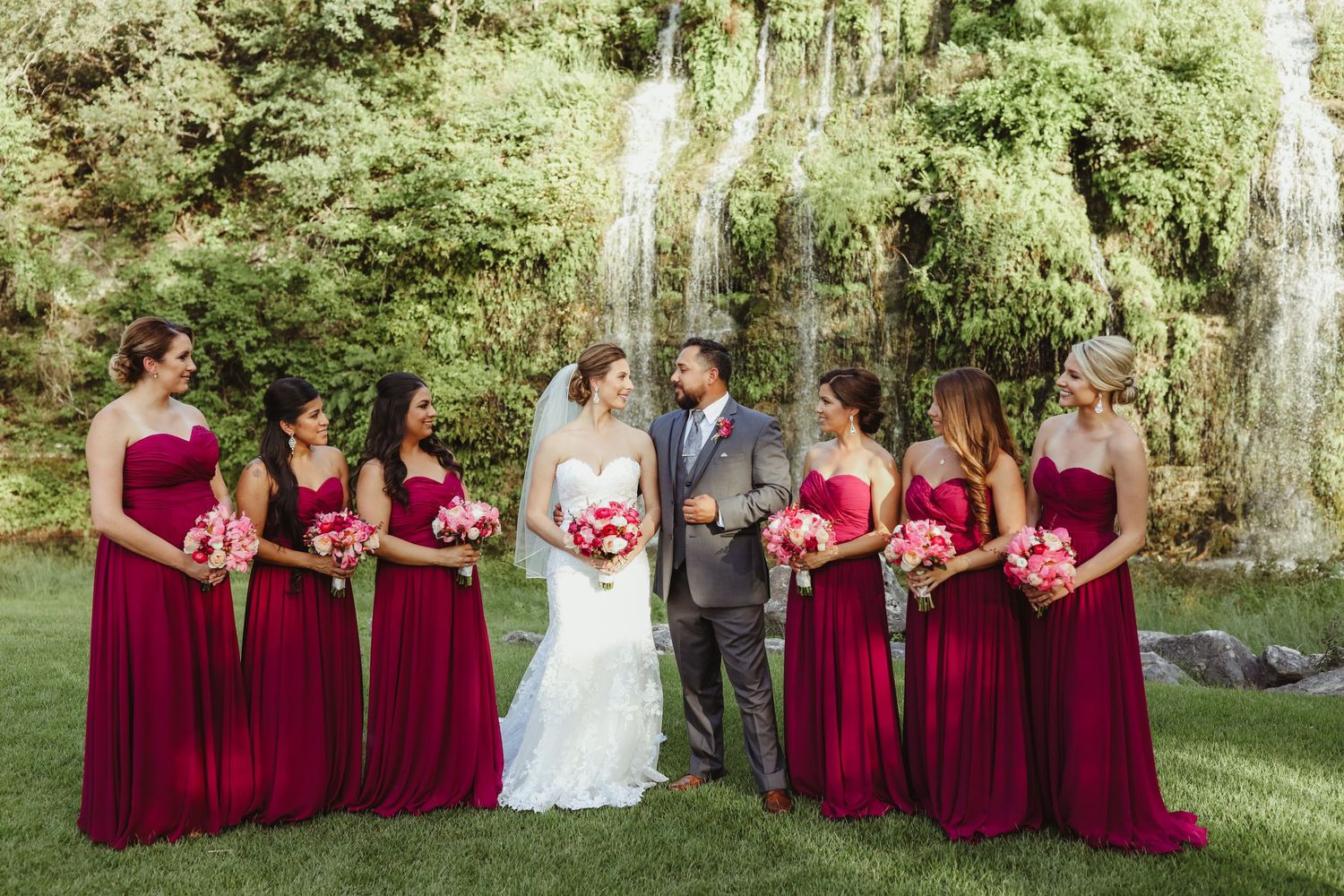 Groom with bridesmaids in red dresses holding pink bouquets near waterfall backdrop.