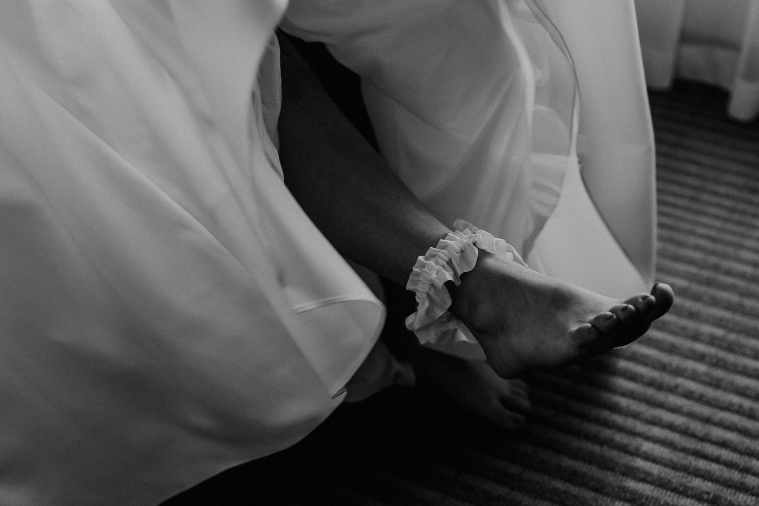 Close-up black and white photo of bride wearing a garter on her ankle beneath wedding dress.