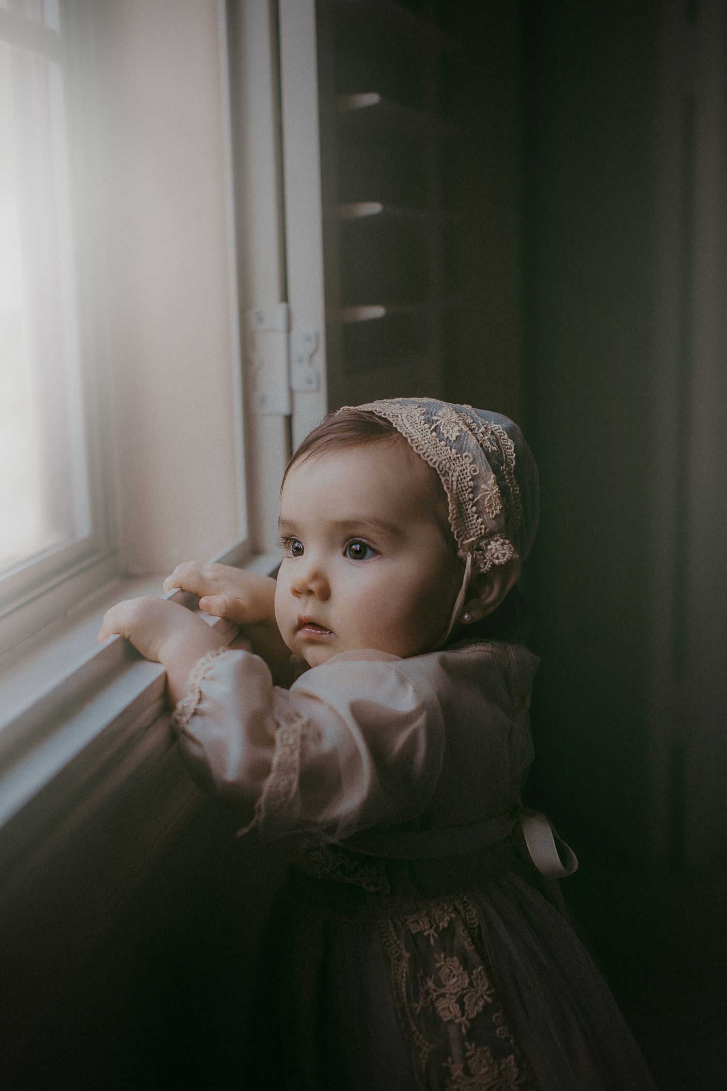 A small child in vintage dress and bonnet gazes pensively through a window in moody lighting.