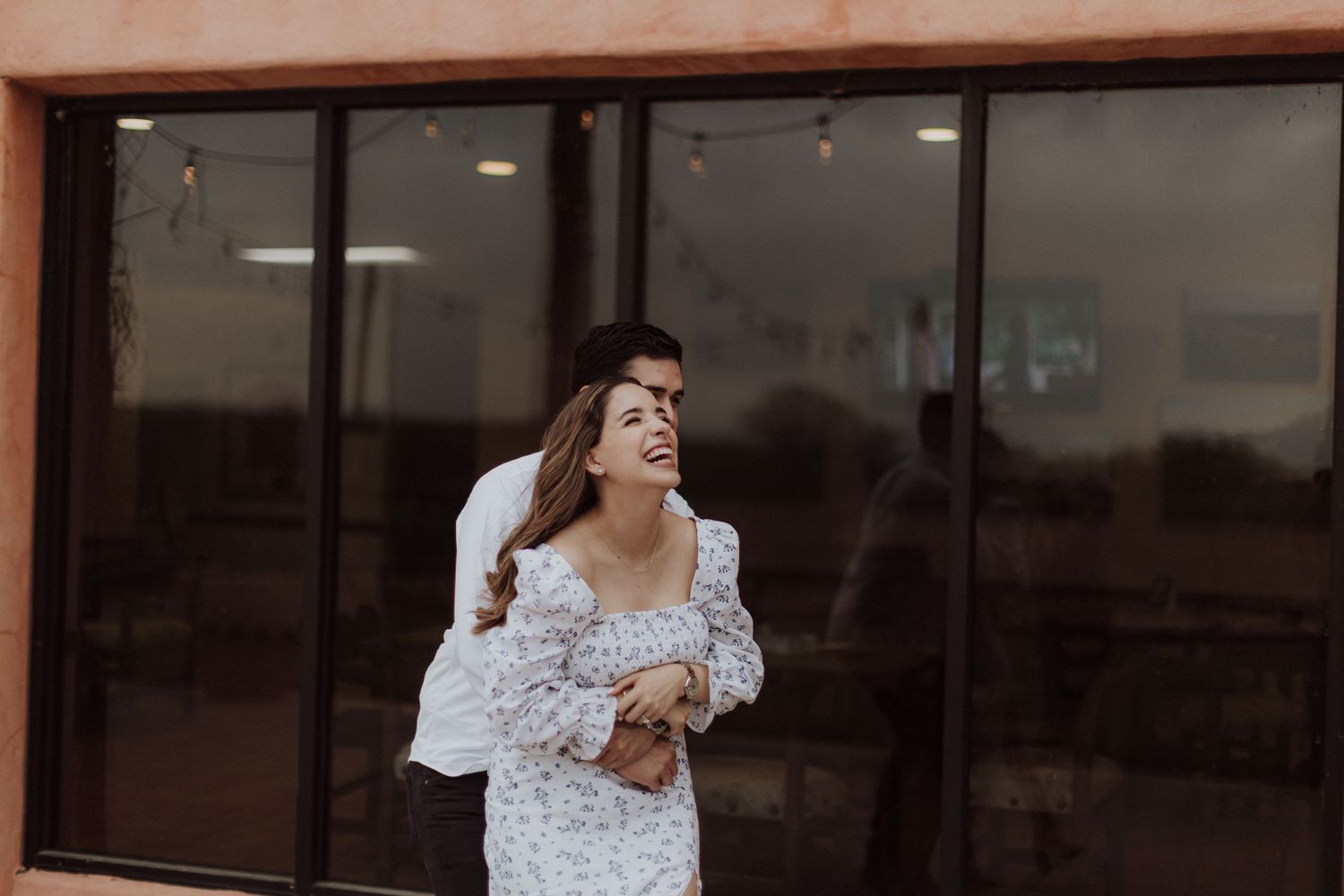 A laughing woman in a floral dress stands outside a building with large windows.