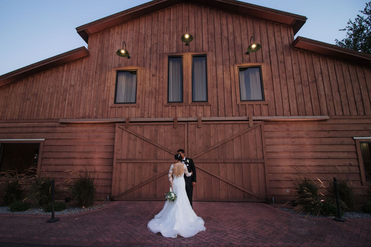 Bride and groom embracing at dusk in front of illuminated rustic red barn.