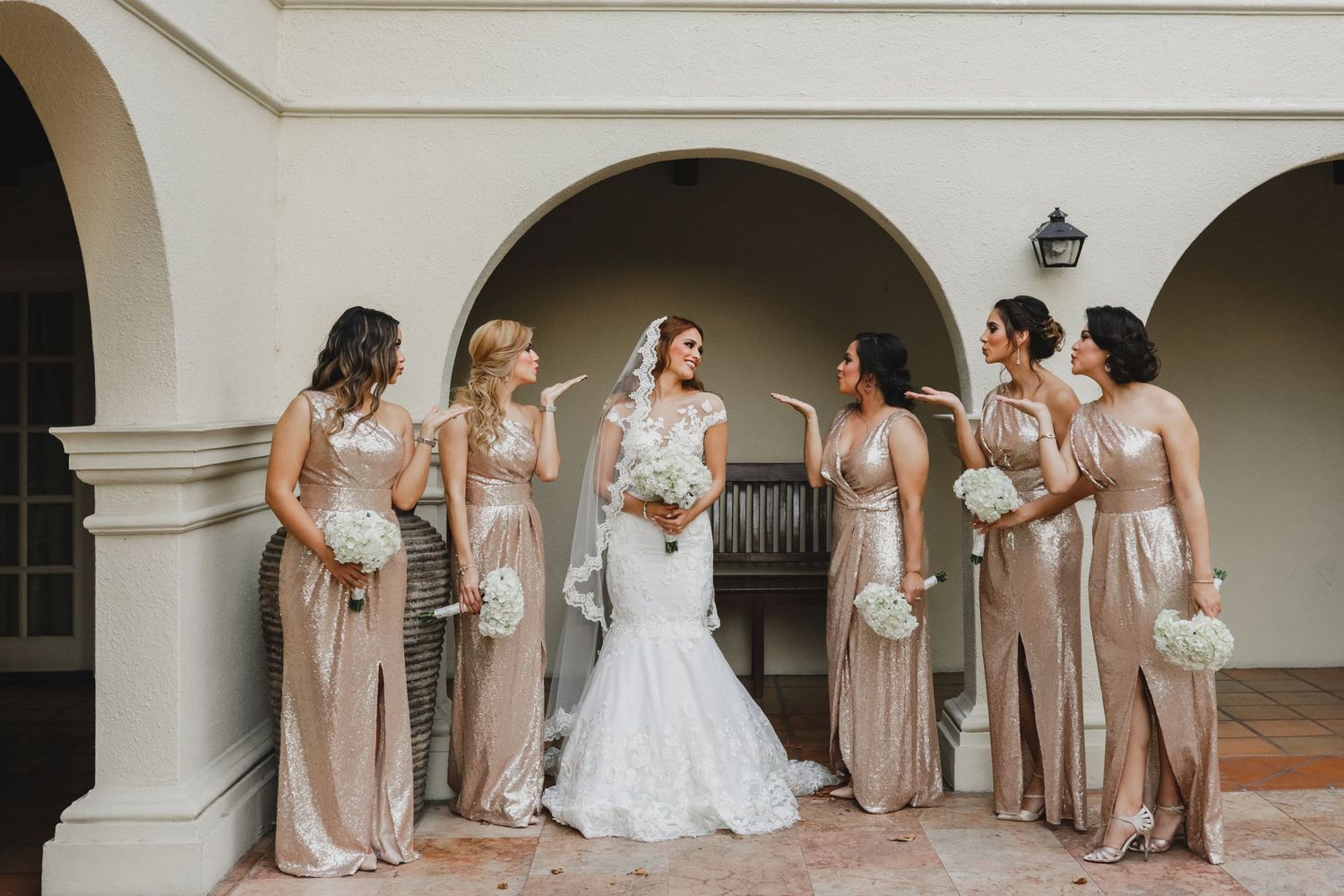 Bride in white gown surrounded by bridesmaids in champagne dresses holding white bouquets.