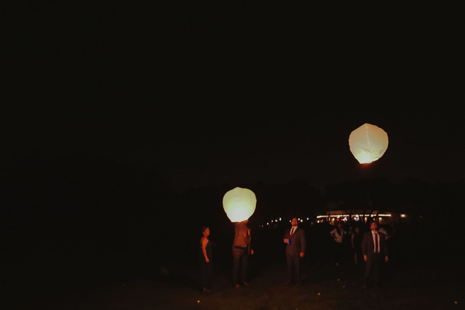 Wedding guests release glowing white sky lanterns into dark night sky outdoors.