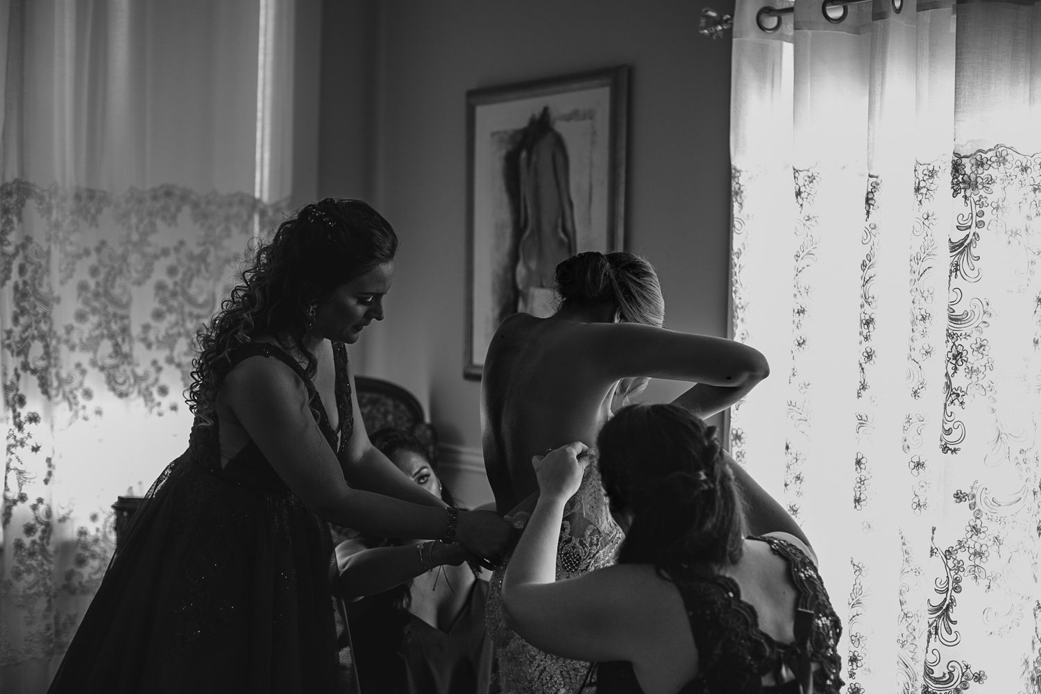 Black and white photo of bridesmaids helping bride into wedding dress near a bright window.