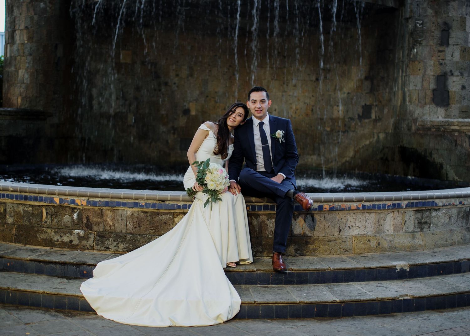 Bride and groom pose together at stone fountain in romantic outdoor wedding portrait.