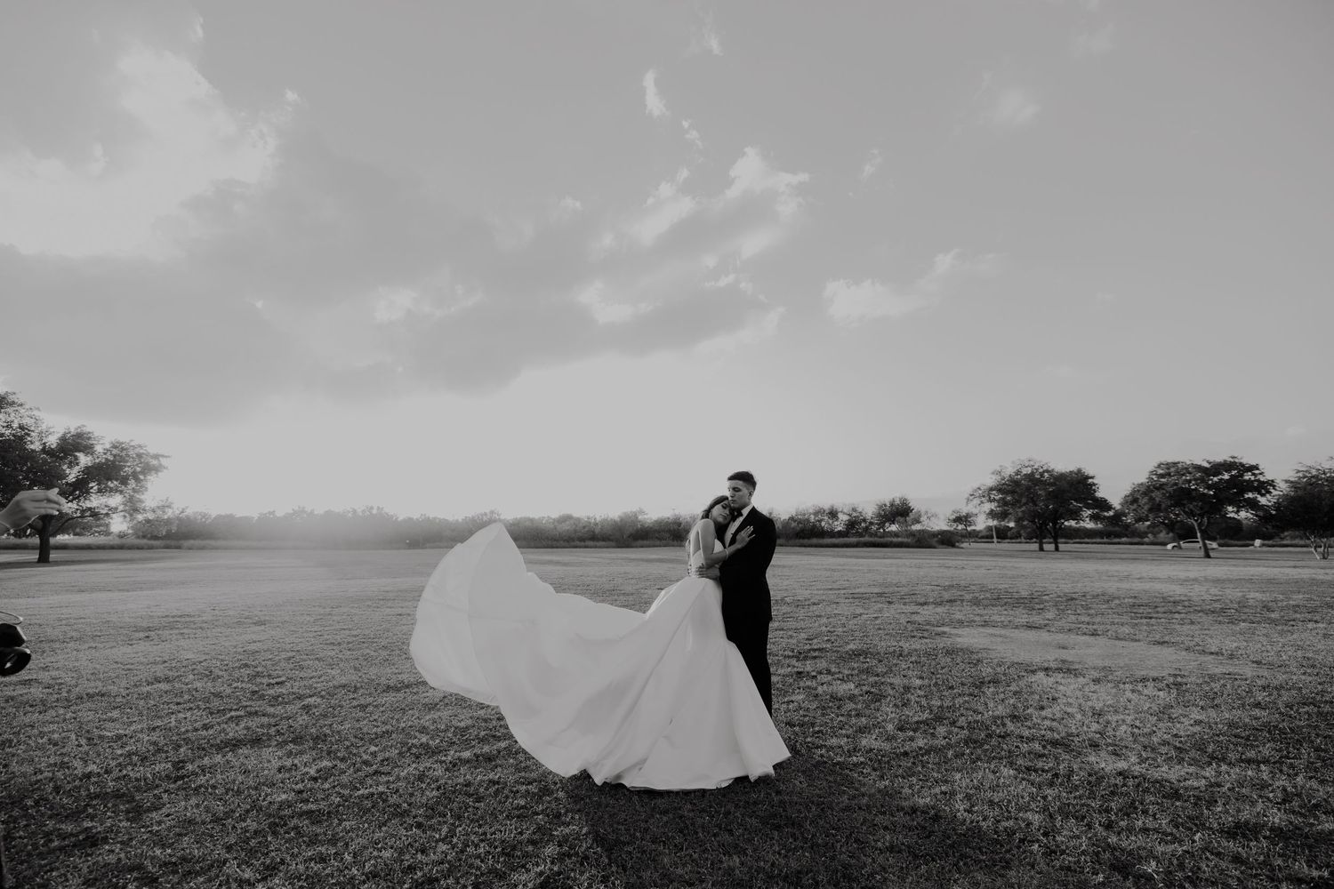 A newlywed couple embraces as the bride's dress billows dramatically in the wind on an open field.
