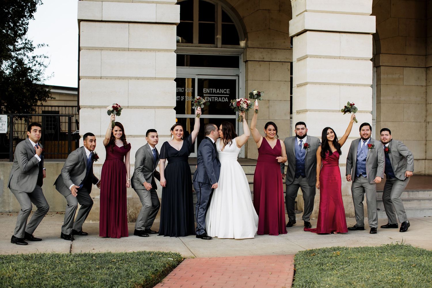 Wedding party posing energetically outside venue entrance in burgundy and gray formal attire.