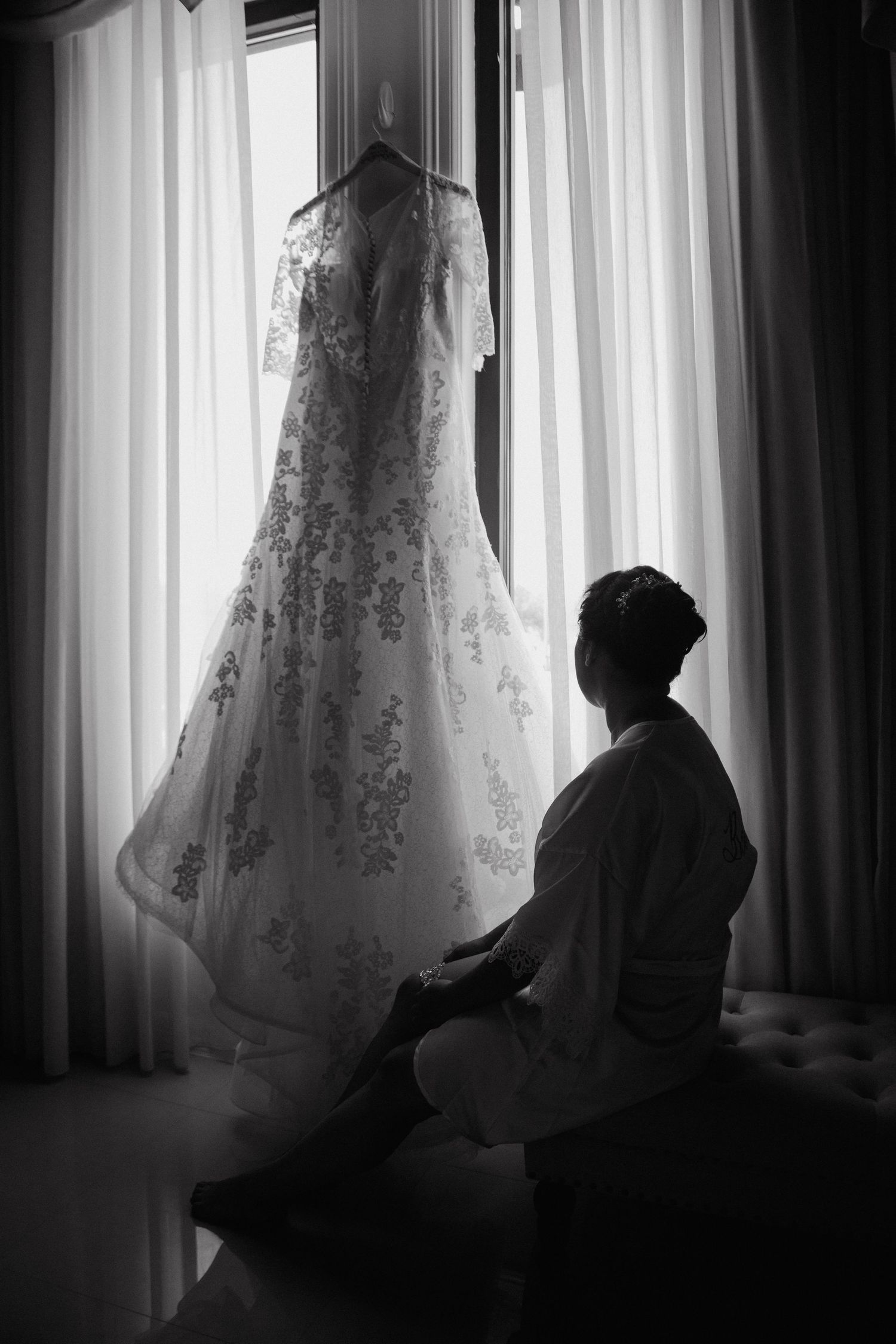 Bride silhouetted against window gazing at lace wedding dress hanging in black and white photo.