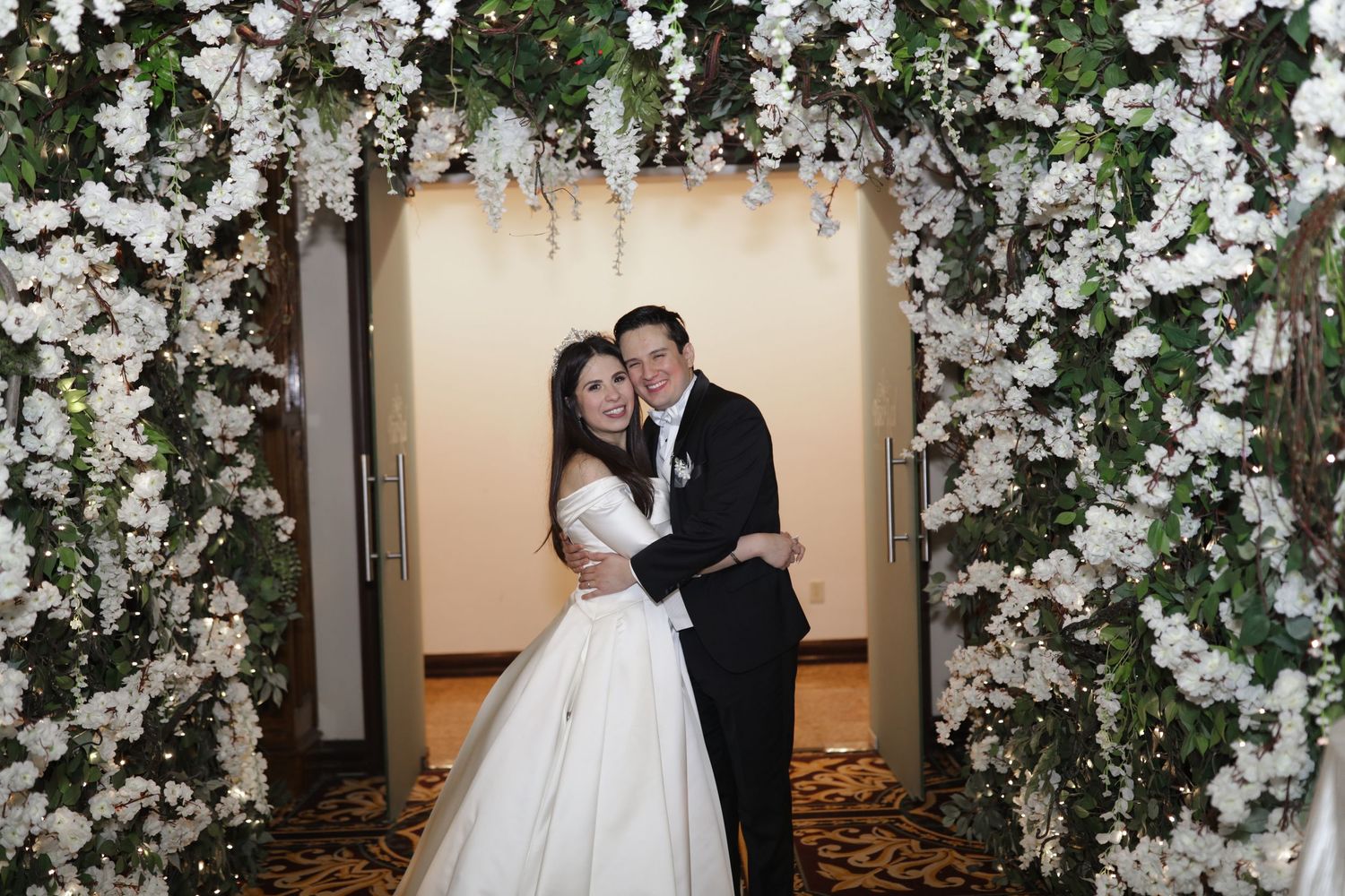 Bride and groom embrace under a floral arch adorned with white flowers at their wedding reception.