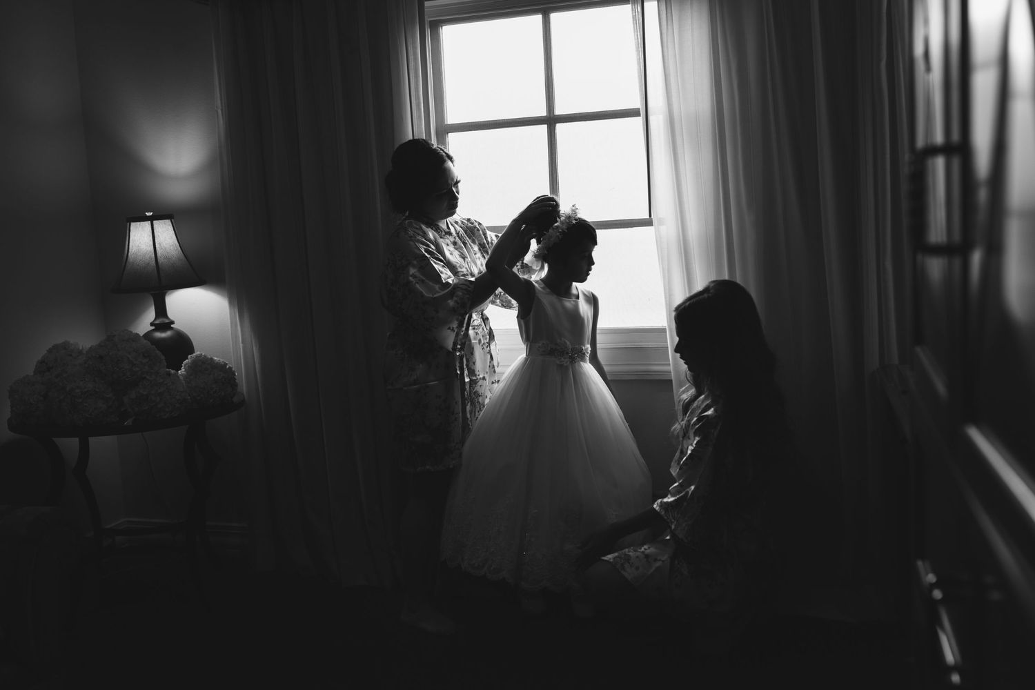 Black and white silhouette of flower girl getting hair done by two women near window.