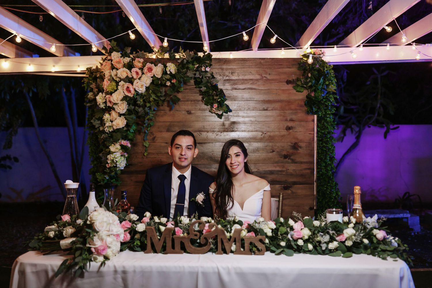 Bride and groom seated at floral-decorated sweetheart table with Mr & Mrs sign and string lights.