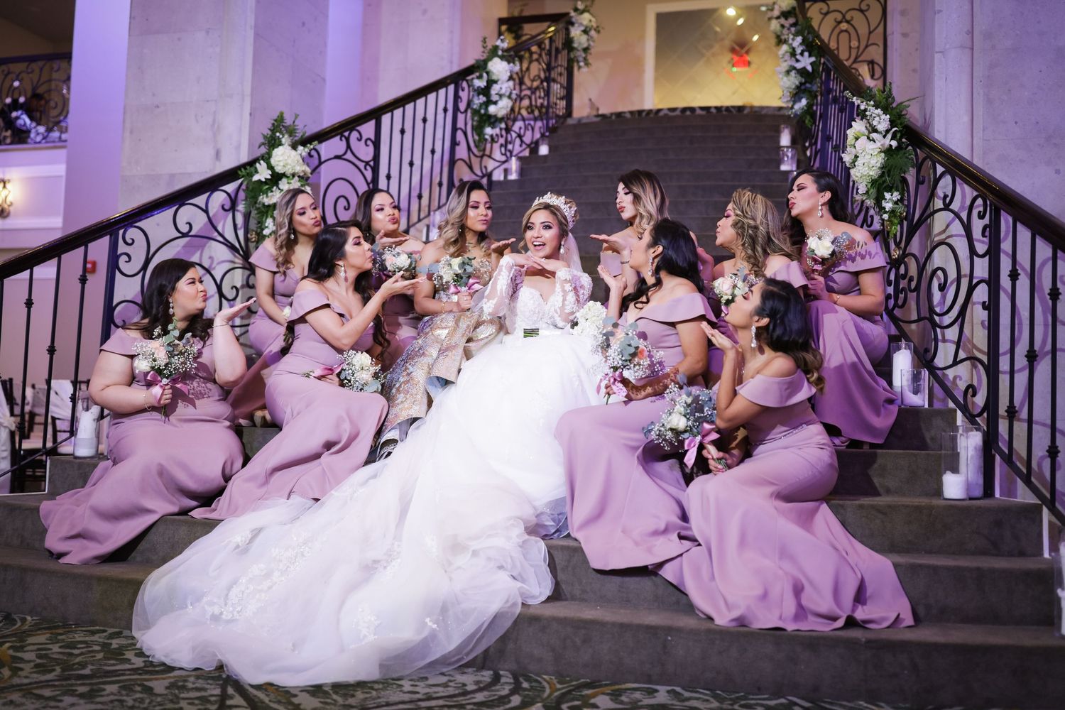 Bride with bridesmaids in lavender mermaid gowns pose on grand staircase decorated with floral arrangements.