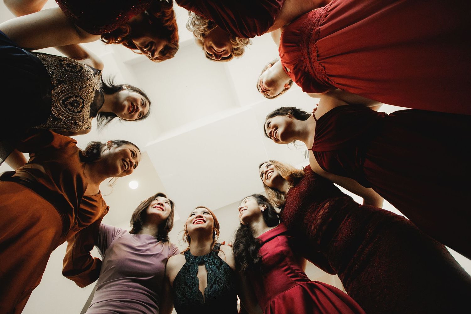 Wedding party forms joyful circle huddle photographed from below showing smiling faces and colorful dresses.