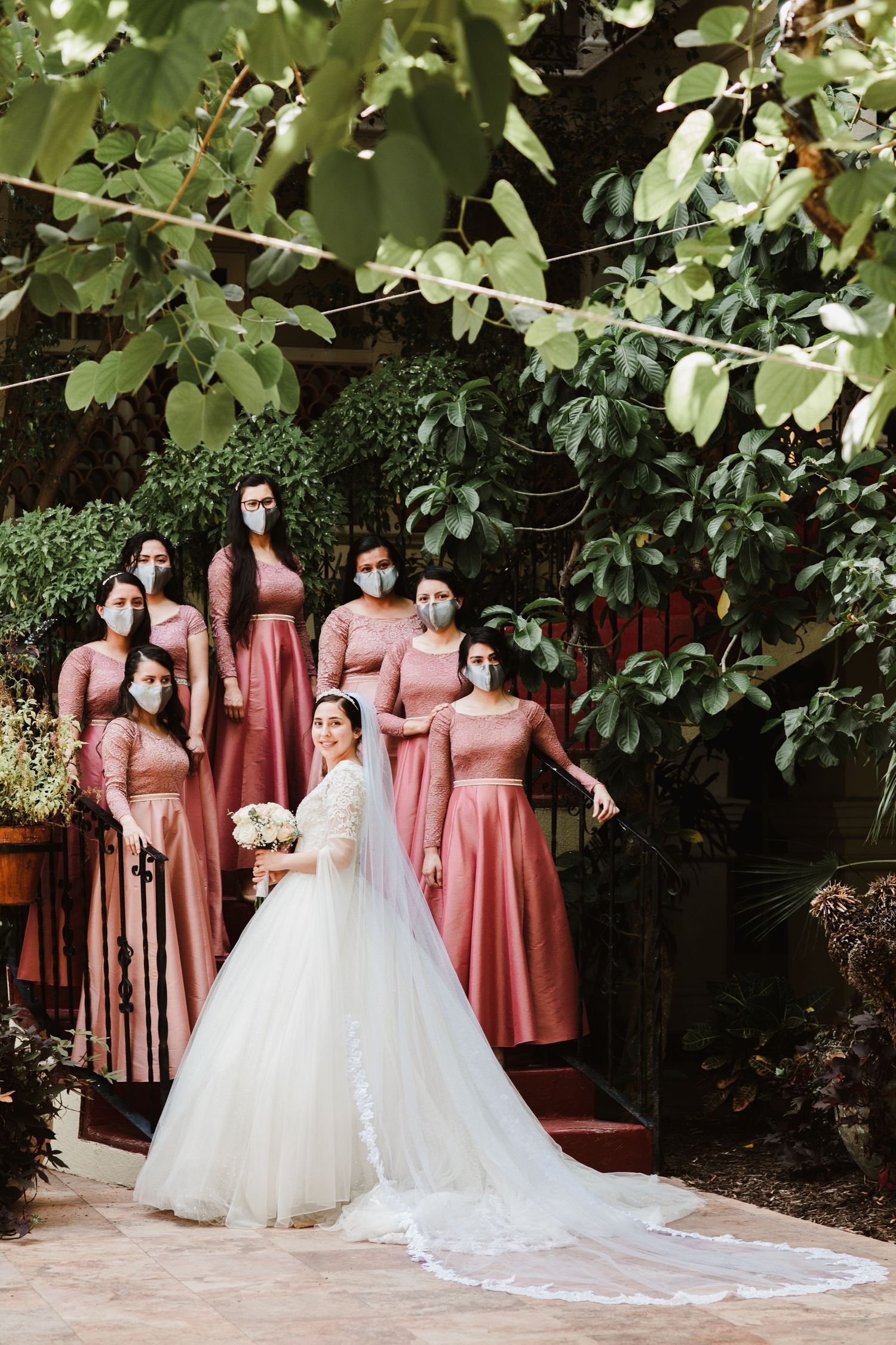 Bride poses with bridesmaids in pink dresses and face masks in a garden setting.