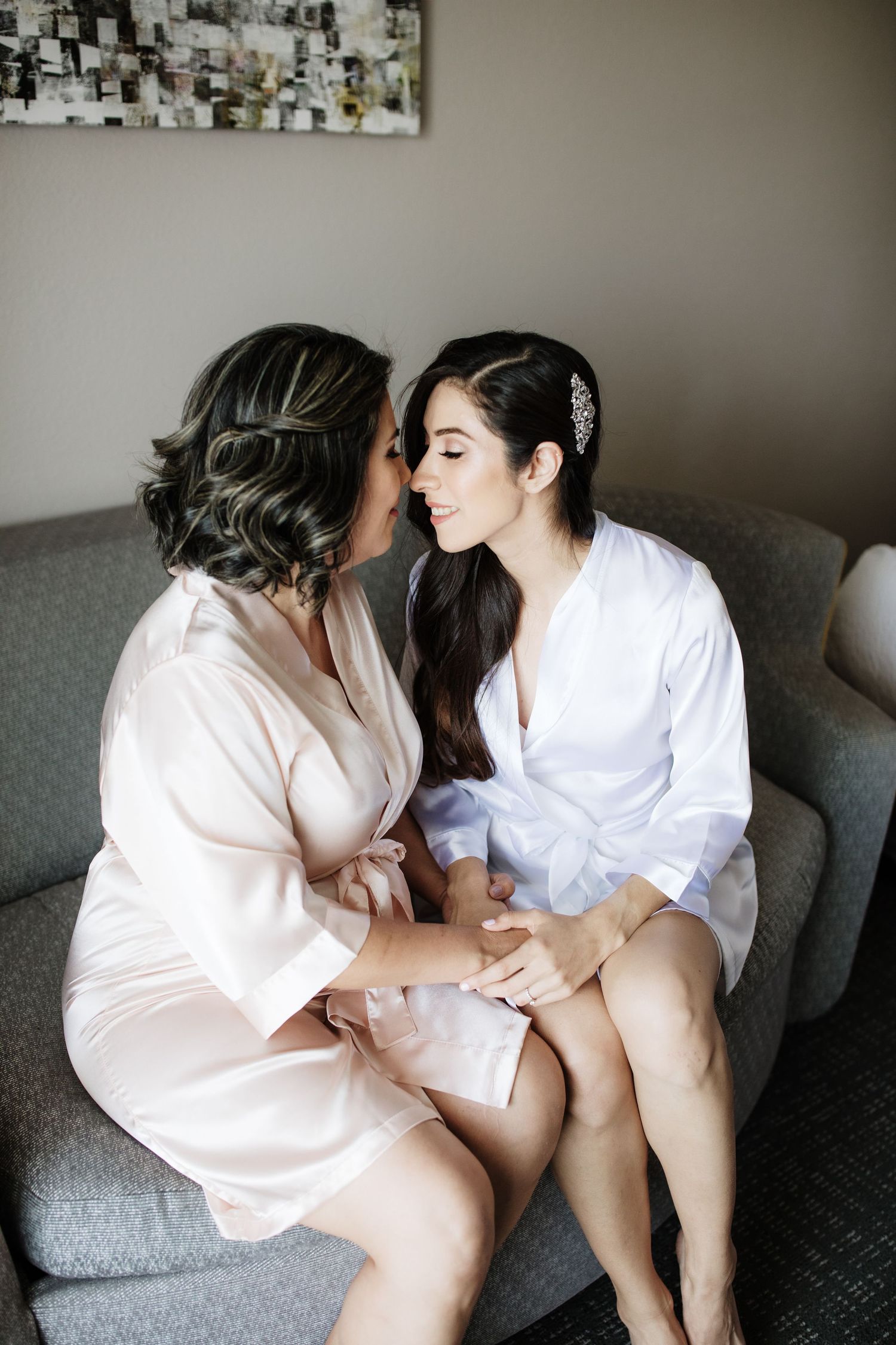 Two women in silk robes share an intimate moment sitting together on a gray sofa during bridal wedding preparations.