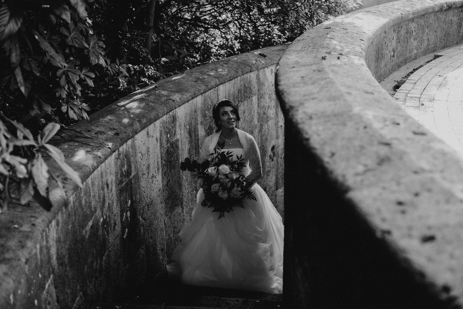 Bride holding a bouquet standing between curved stone walls in black and white photo.