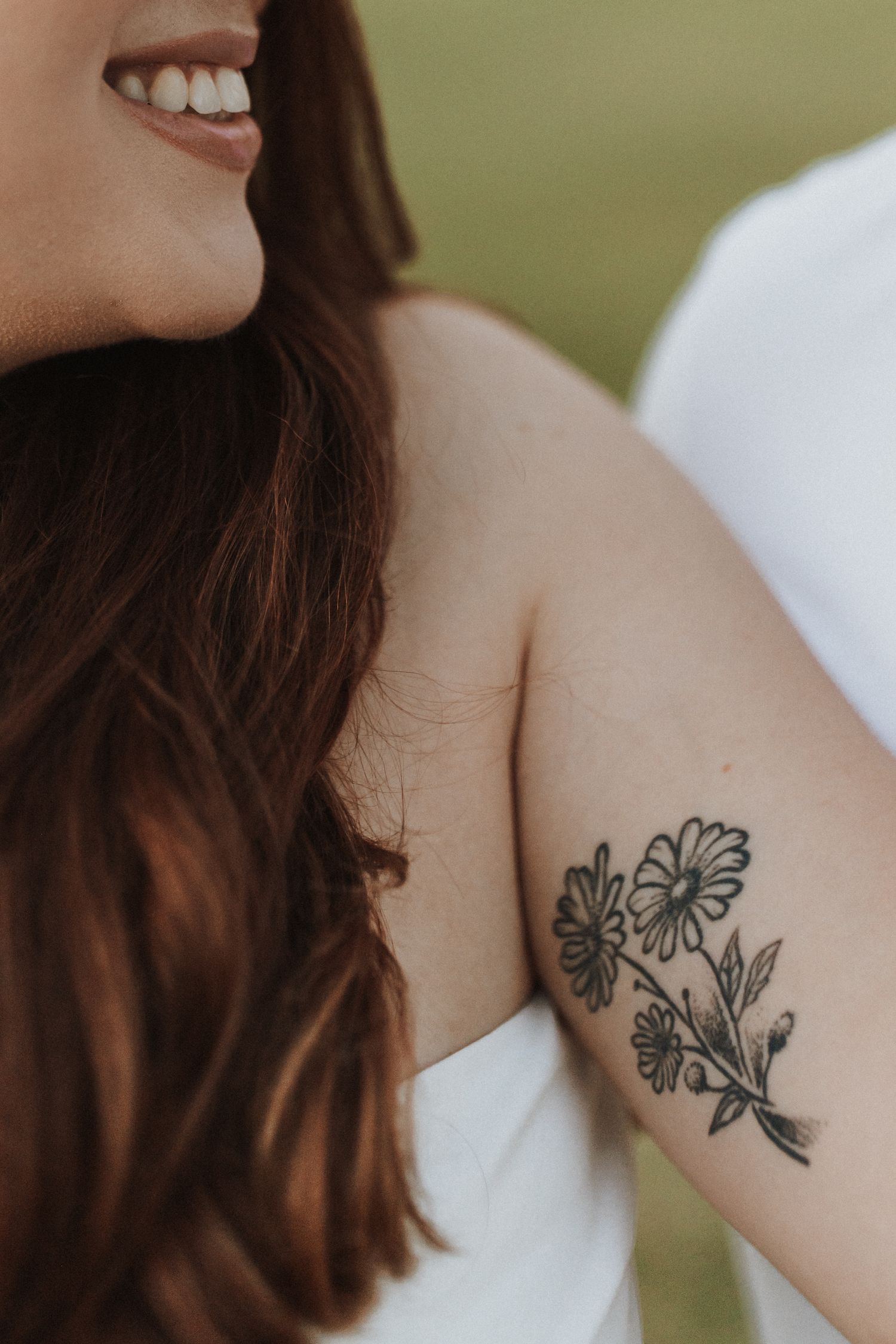 Smiling woman with long brown hair wearing white dress showing floral daisy tattoo on upper arm.