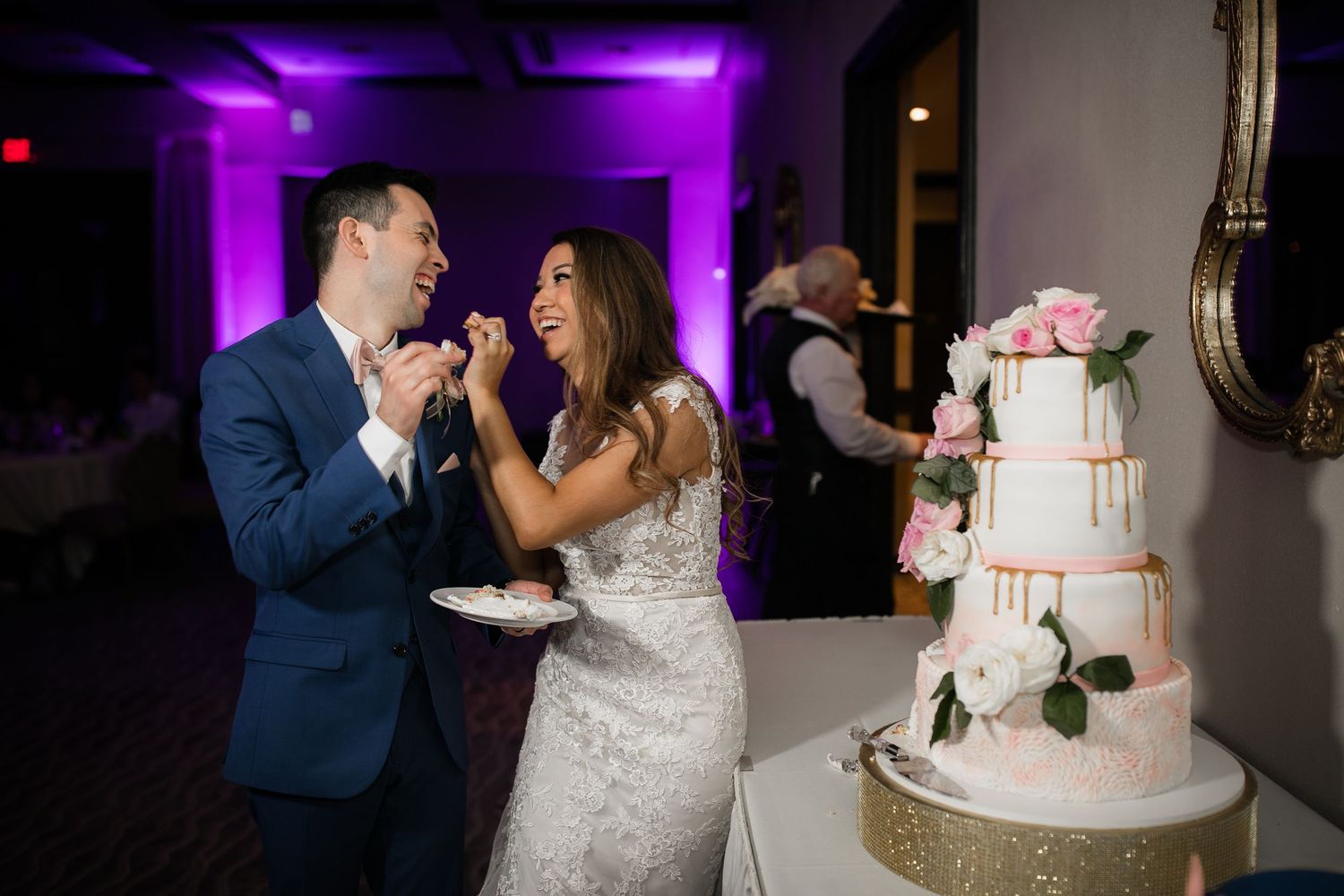 Bride and groom laugh joyfully while cutting their elegant four-tier white wedding cake adorned with pink roses.