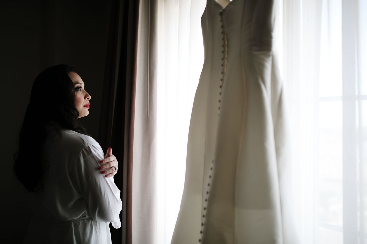 Bride in white robe gazing at her wedding dress hanging by the window in soft natural light.